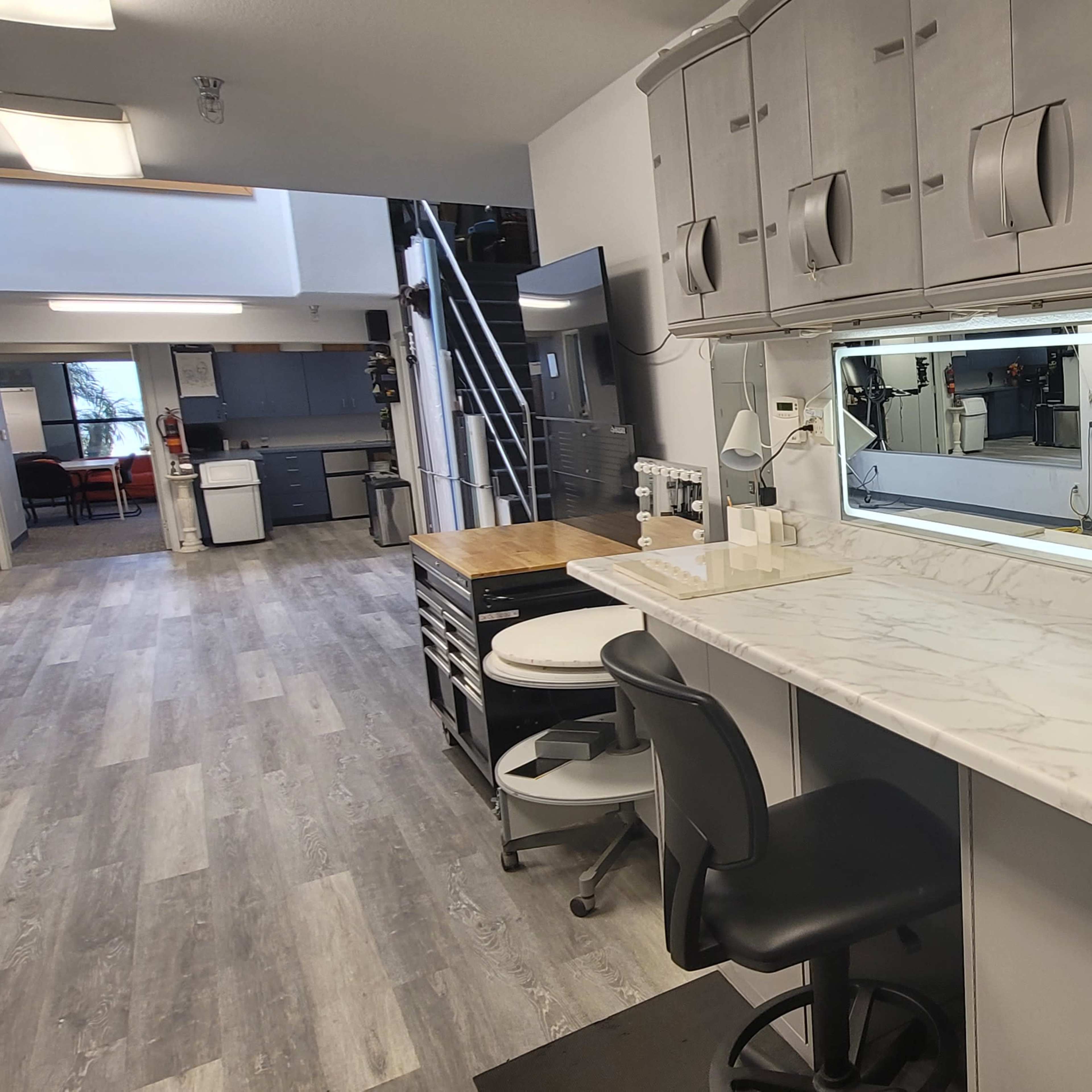 A modern workspace with a long marble countertop, two black chairs, and overhead storage cabinets, illuminated by a backlit mirror.