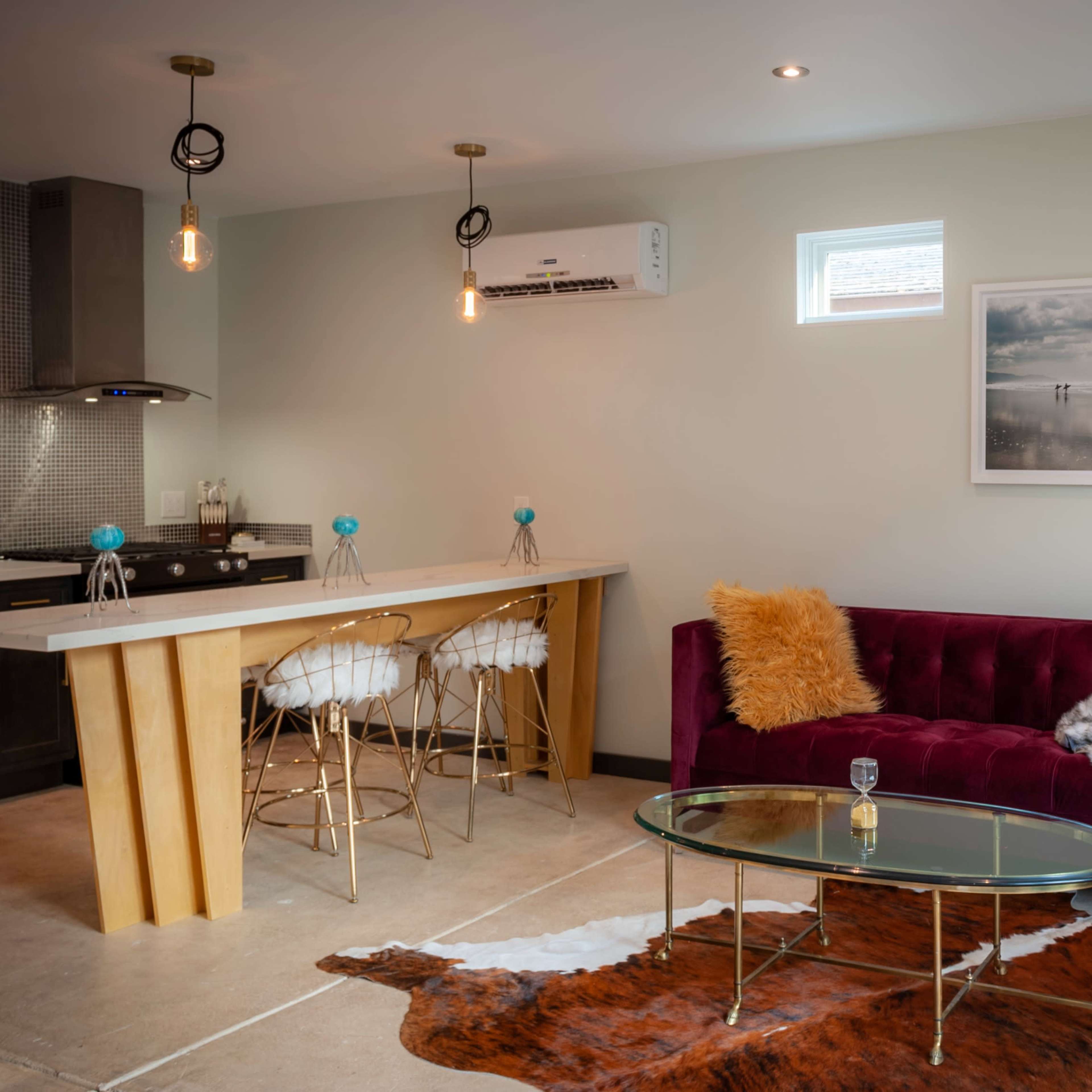 A modern living space featuring a purple velvet sofa, a glass coffee table on a cowhide rug, and a kitchen area in the background with a bar and stools.