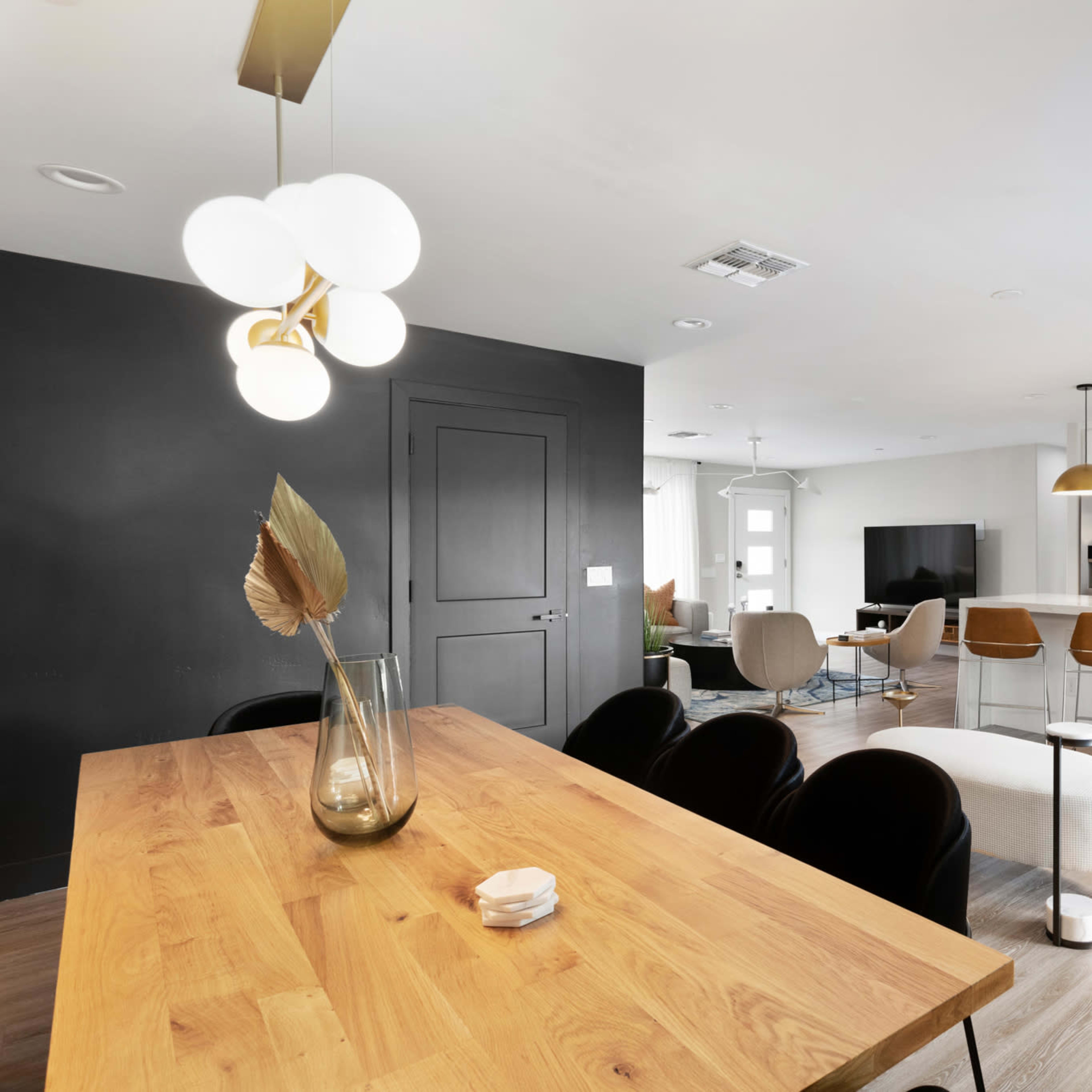 A modern open-concept living space featuring a dining area with a wooden table, a black accent wall, and a kitchen equipped with white cabinetry and gold pendant lights.