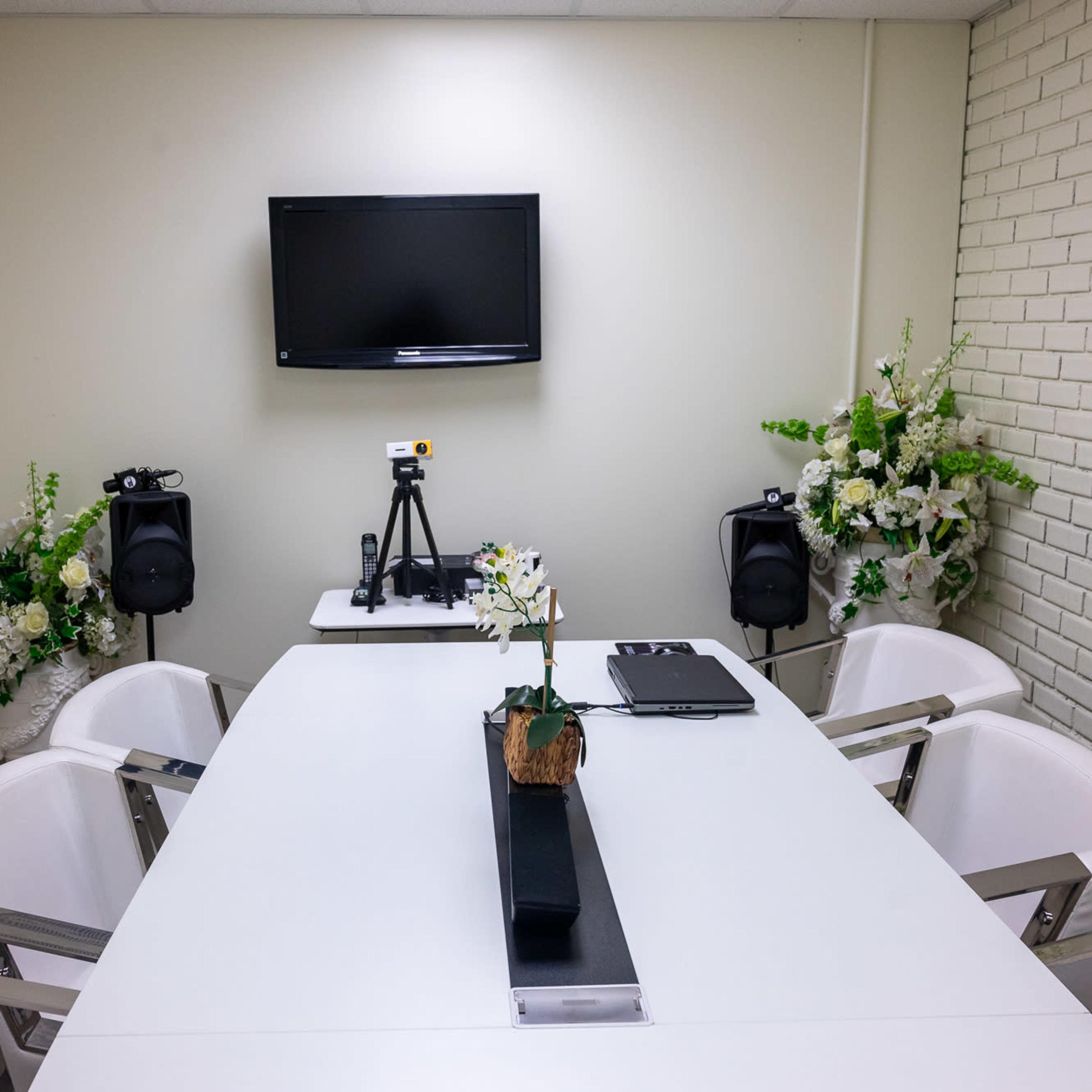 A conference room features a long white table surrounded by six white chairs, with a mounted television and two speakers at opposite ends, and floral arrangements on either side.
