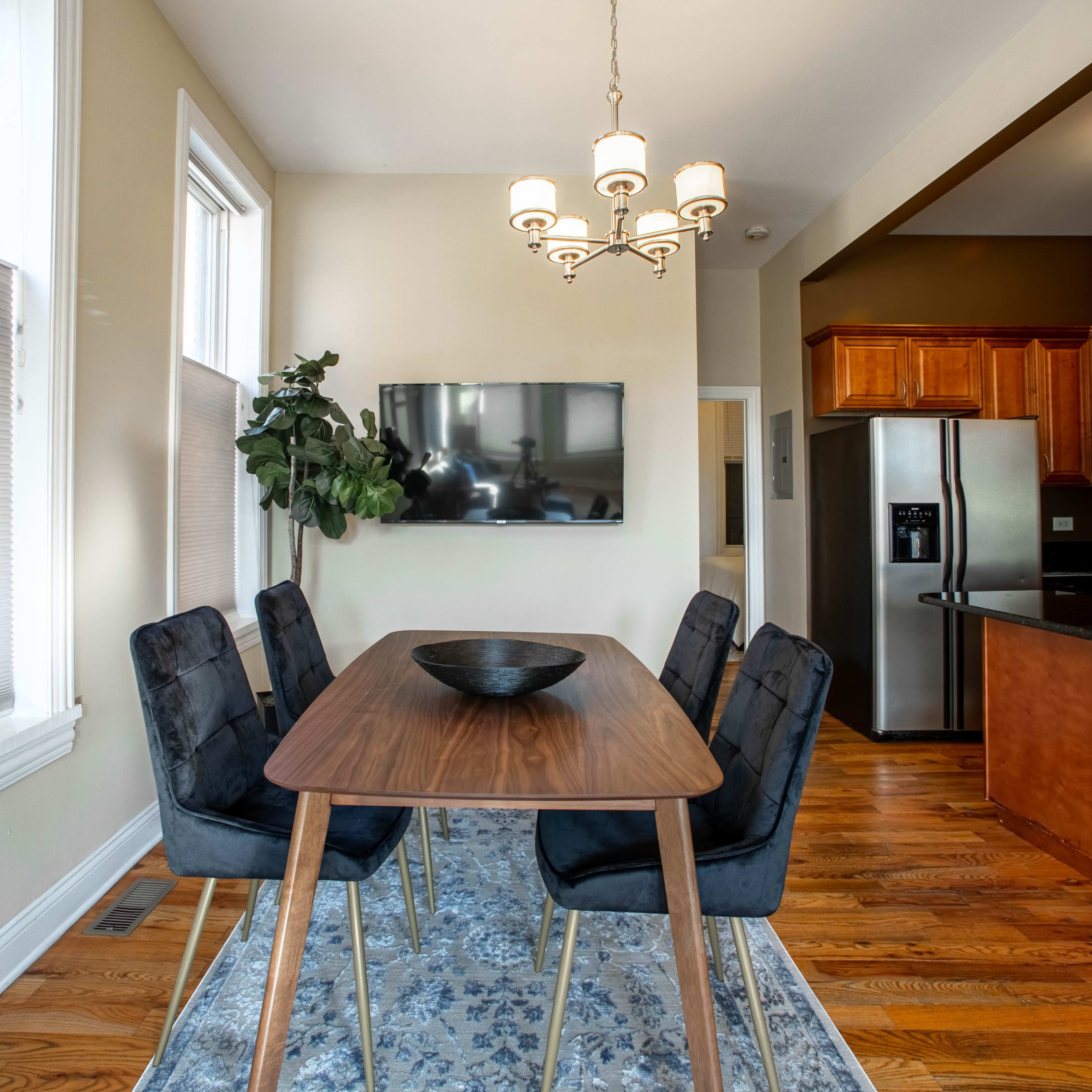 A dining area features a wooden table surrounded by four chairs, with a wall-mounted television and a plant nearby, illuminated by a chandelier above.