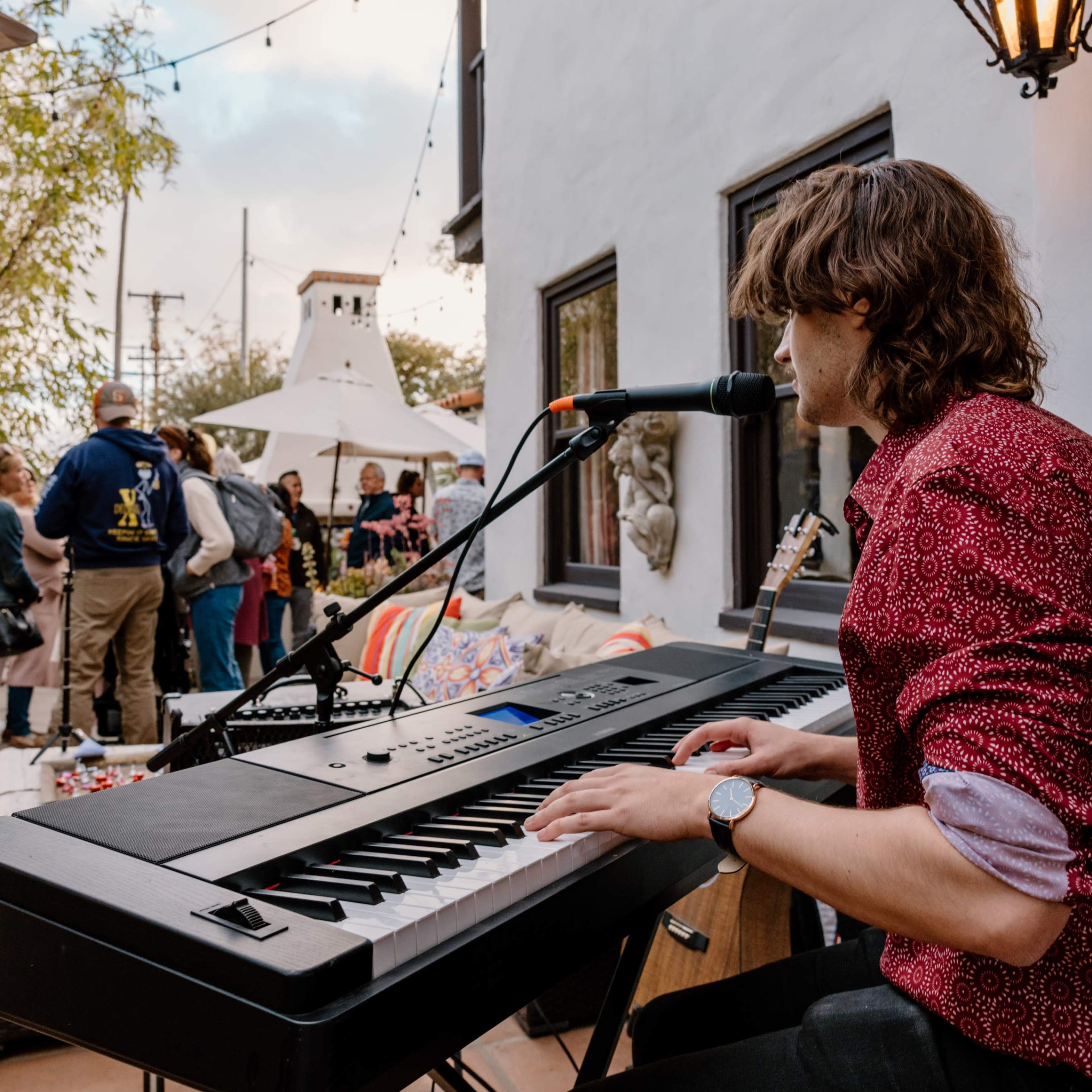 A musician plays a keyboard on an outdoor stage while an audience gathers nearby.