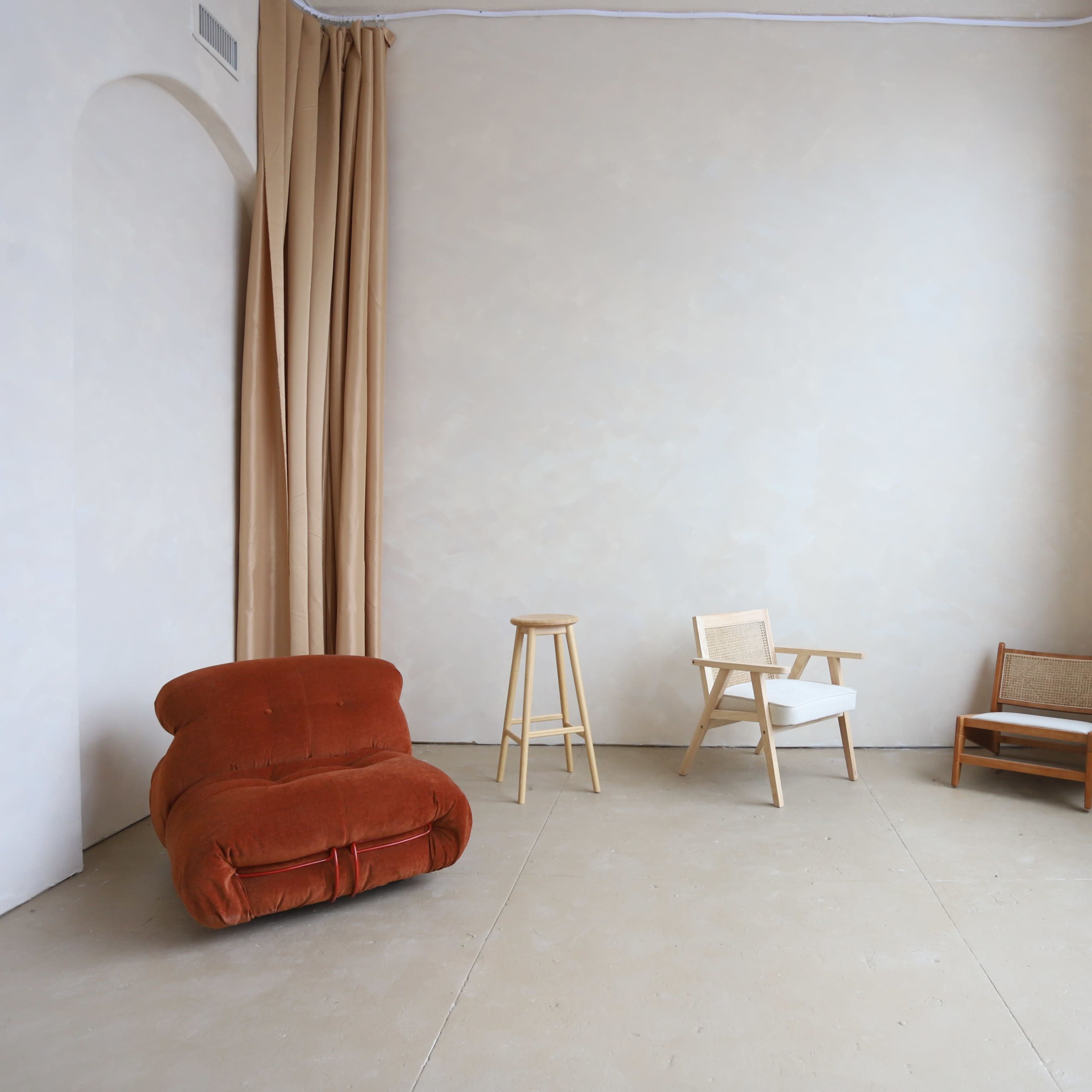 A minimalist interior space featuring an orange upholstered chair, a wooden stool, and two other various styled chairs near a large window.
