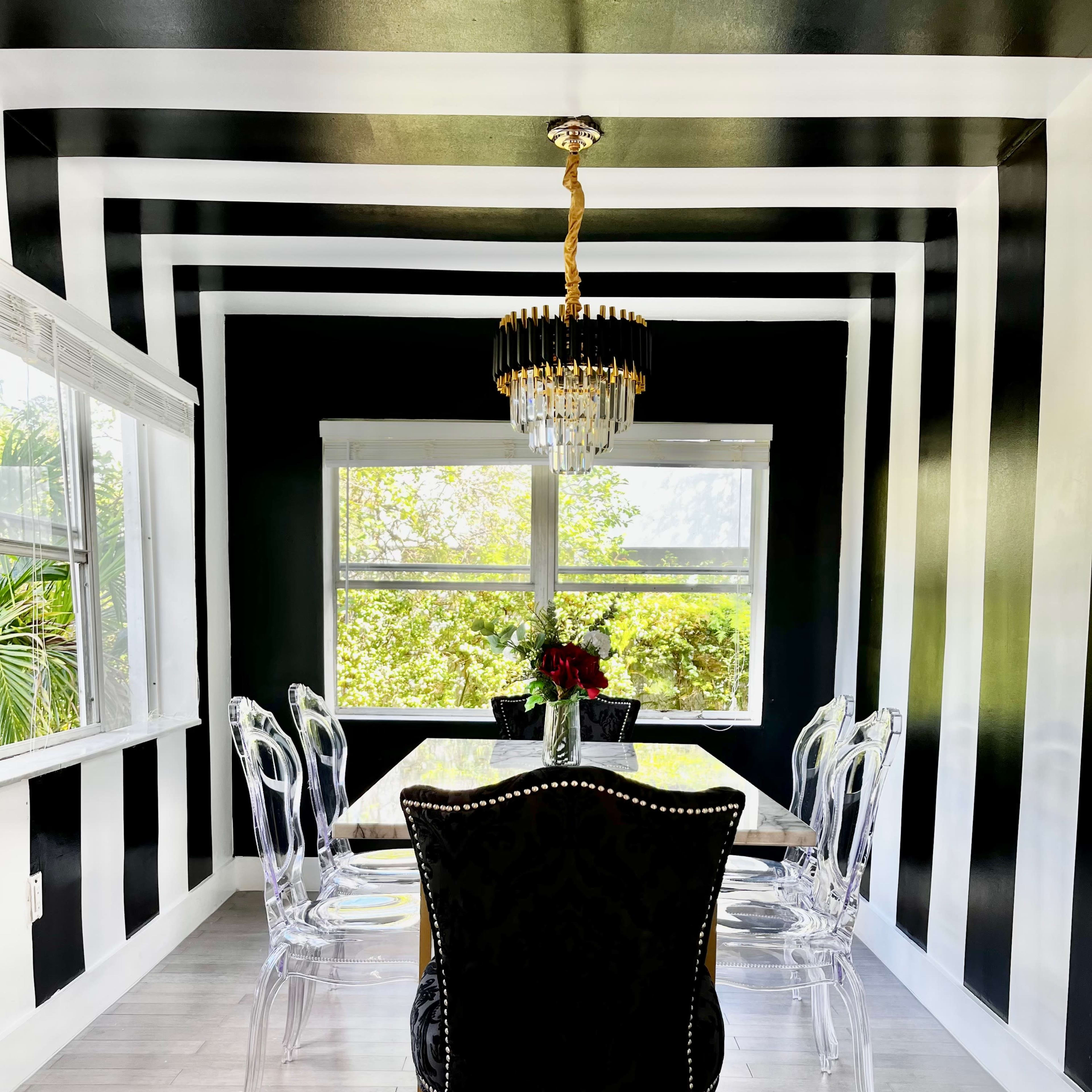 A dining room features a striped black and white wall design, a crystal chandelier, a glass table, and transparent chairs.