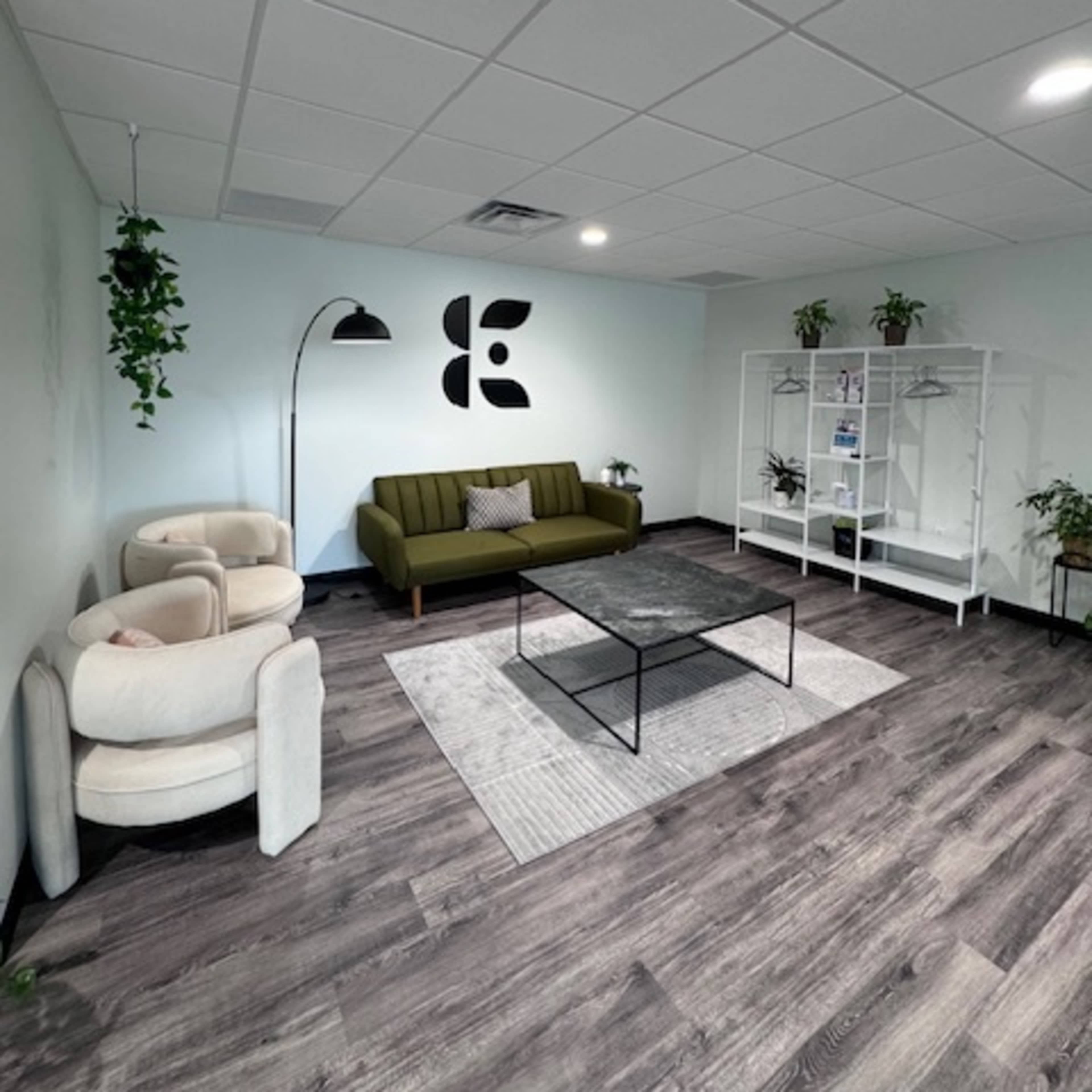 A modern office lounge area with a green sofa, two cream-colored chairs, a black coffee table, and a white bookshelf, all complemented by small plants and a floor lamp.