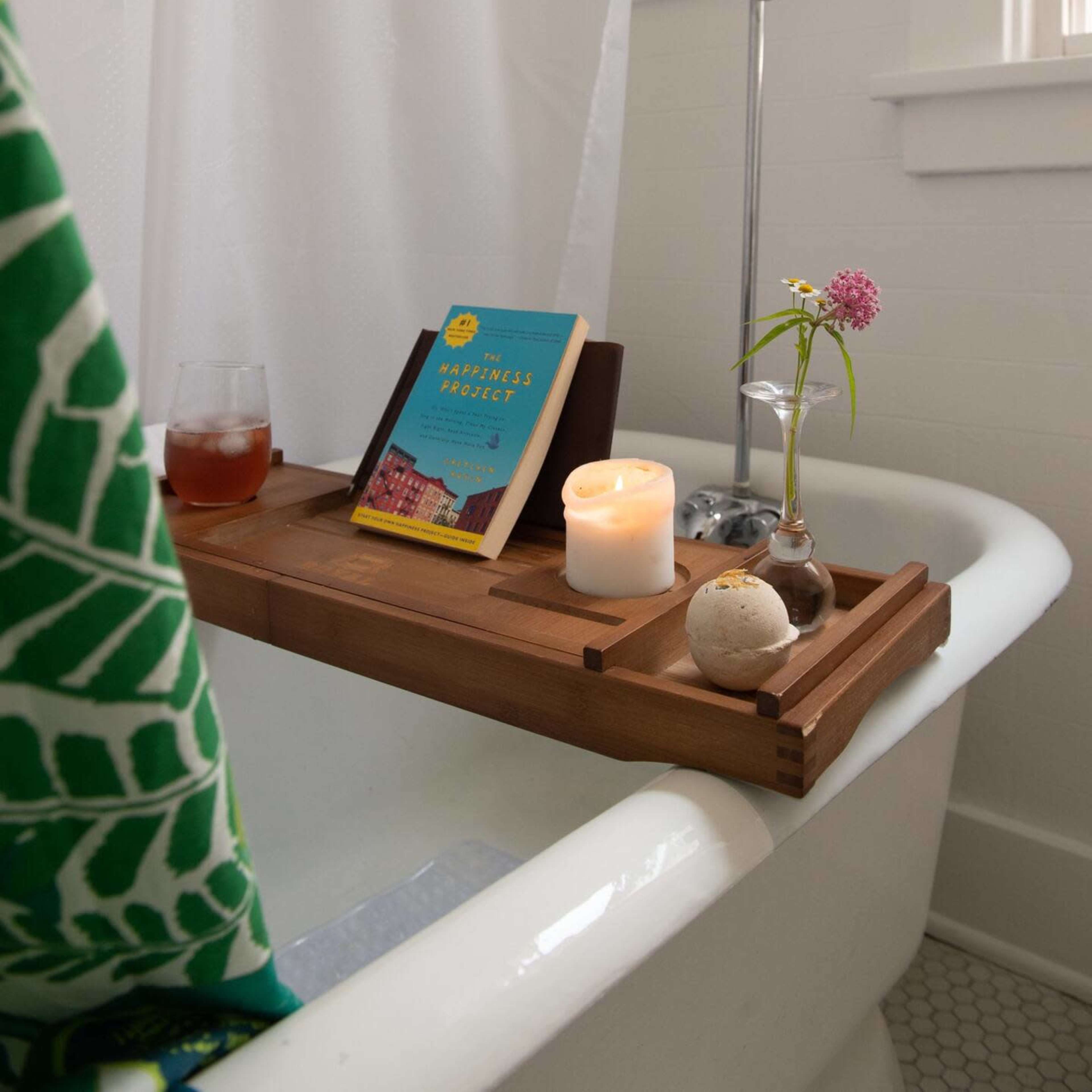 A wooden bath tray holding a book, a candle, a glass of drink, and a small flower vase is placed across the edge of a clawfoot bathtub.