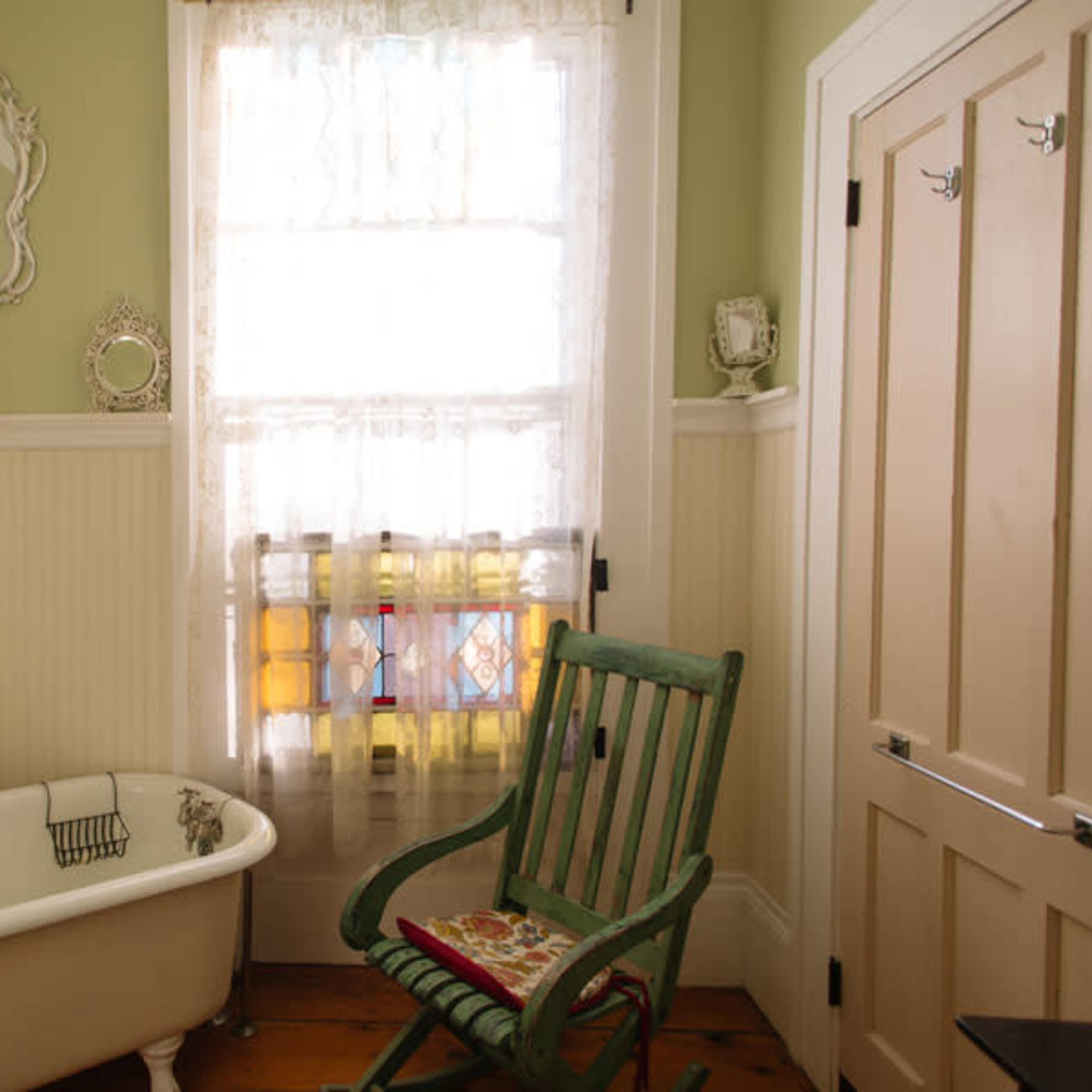 A bathroom featuring a clawfoot bathtub, a green rocking chair, and a window with stained glass.