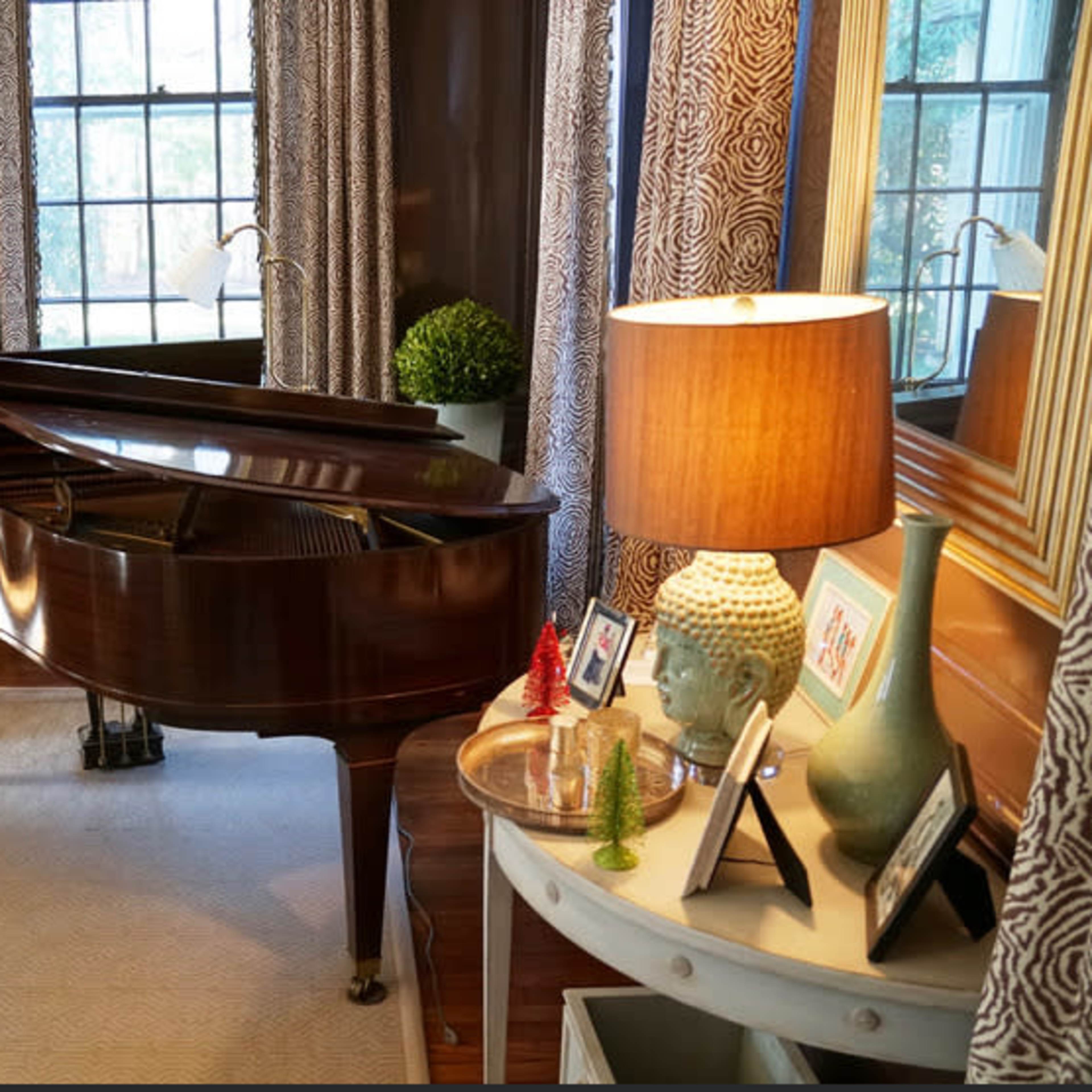 A grand piano sits in a well-lit living room adorned with decorative curtains and furnishings, beside a lamp and a small round table displaying framed photos and a decorative vase.