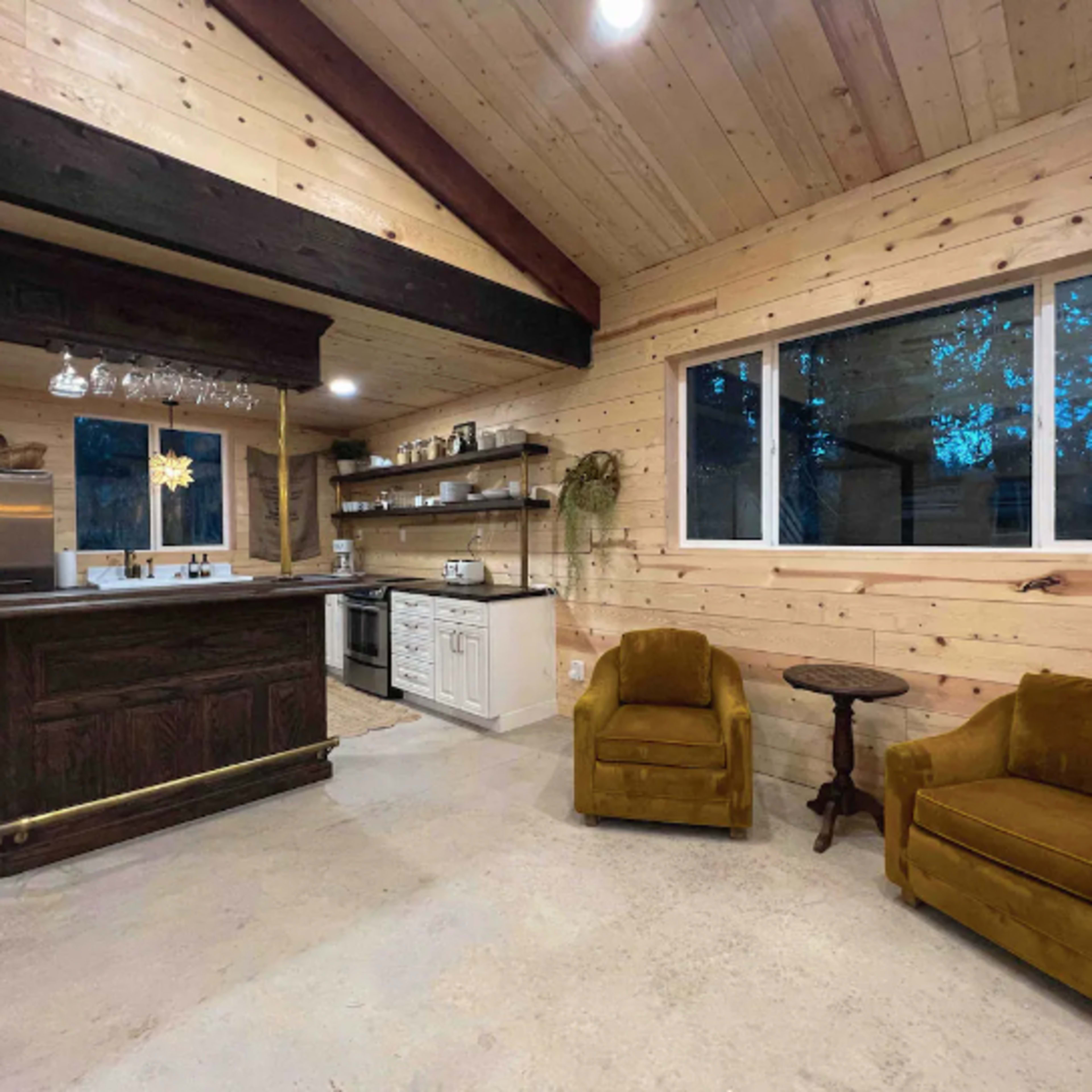 A rustic kitchen and living area featuring wooden walls, a bar with a hanging glass rack, and two yellow chairs beside a small table.