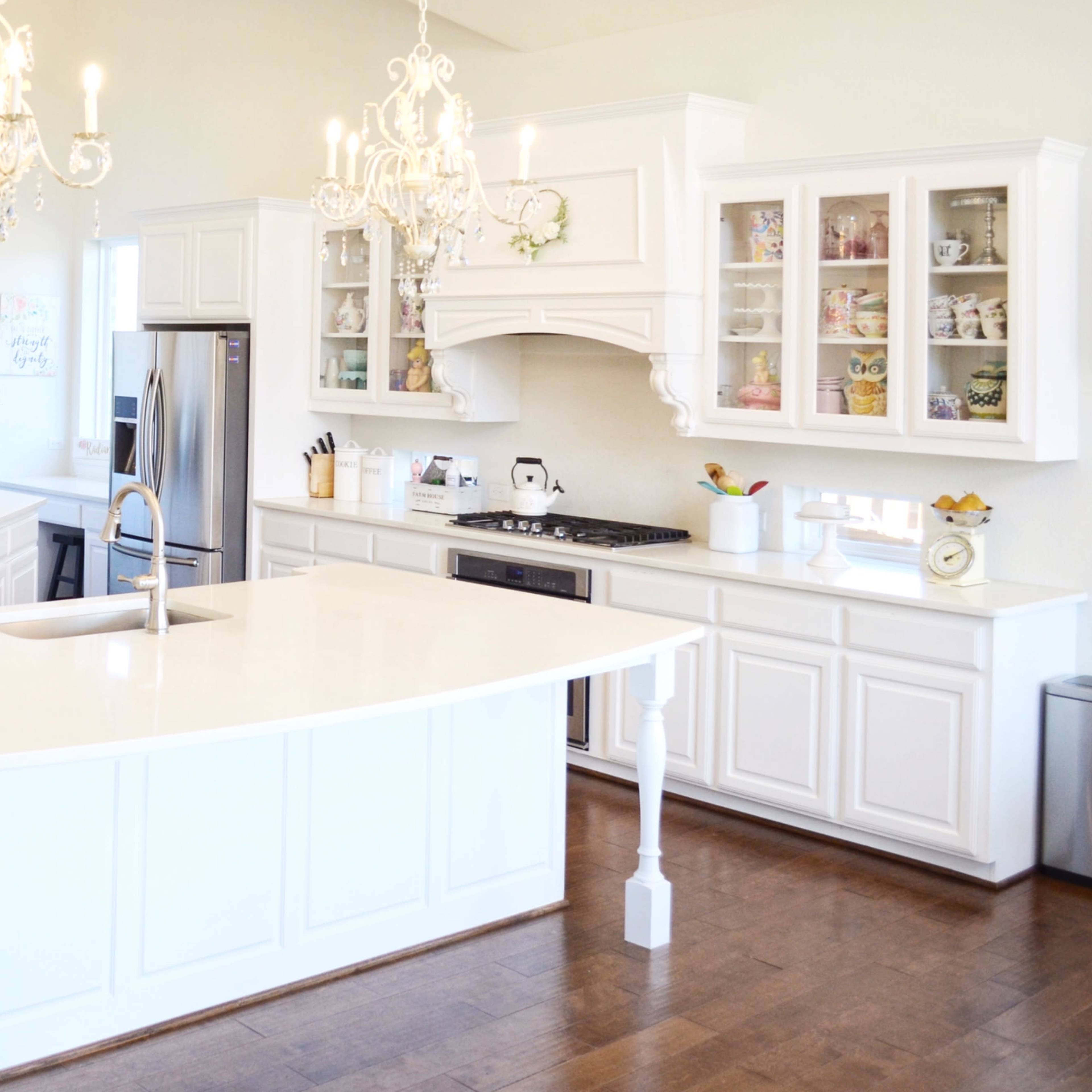 A spacious kitchen features white cabinetry, a large island with a sink, stainless steel appliances, and two chandeliers hanging from the ceiling.