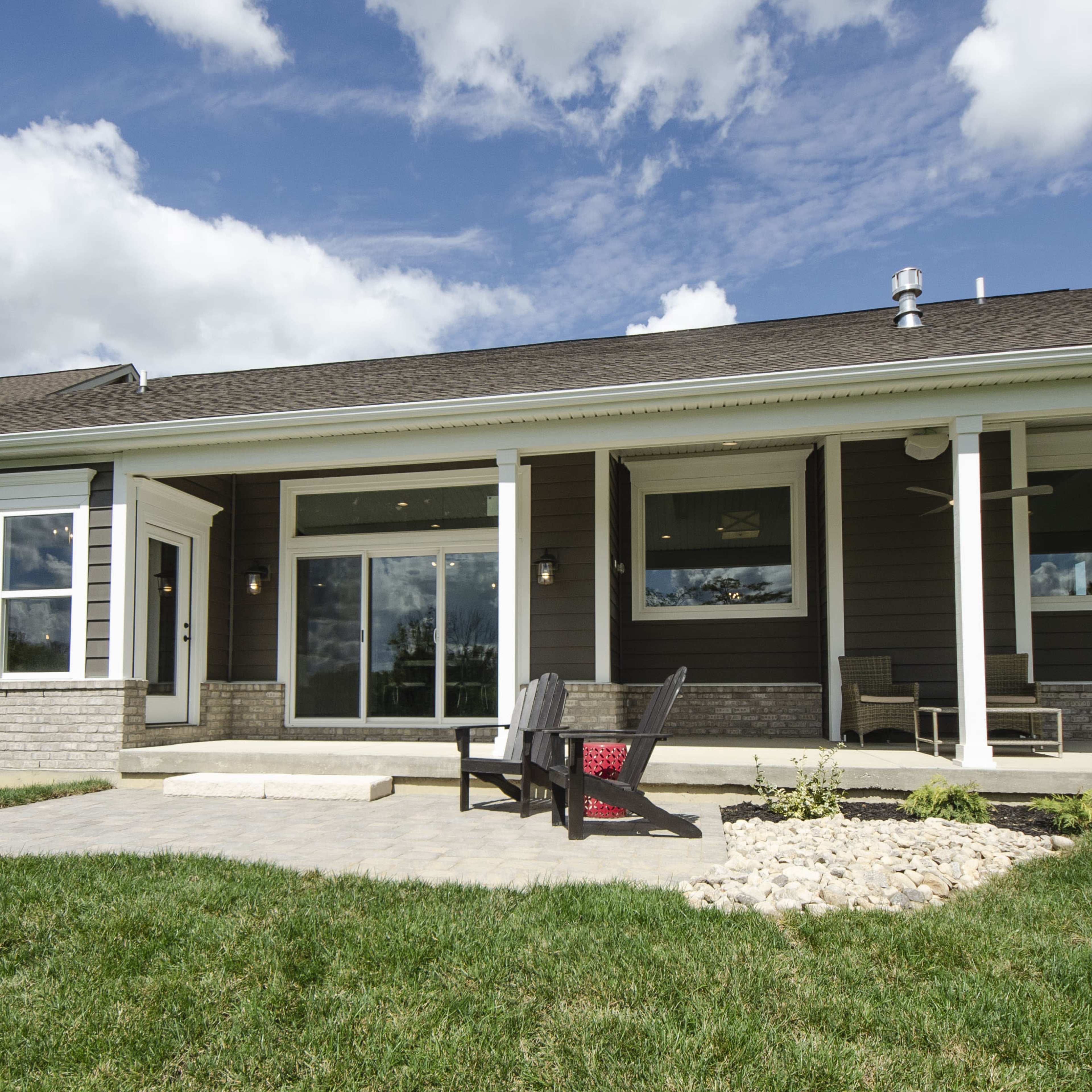A single-story house features a front porch with seating, large windows, and landscaped greenery under a cloudy sky.