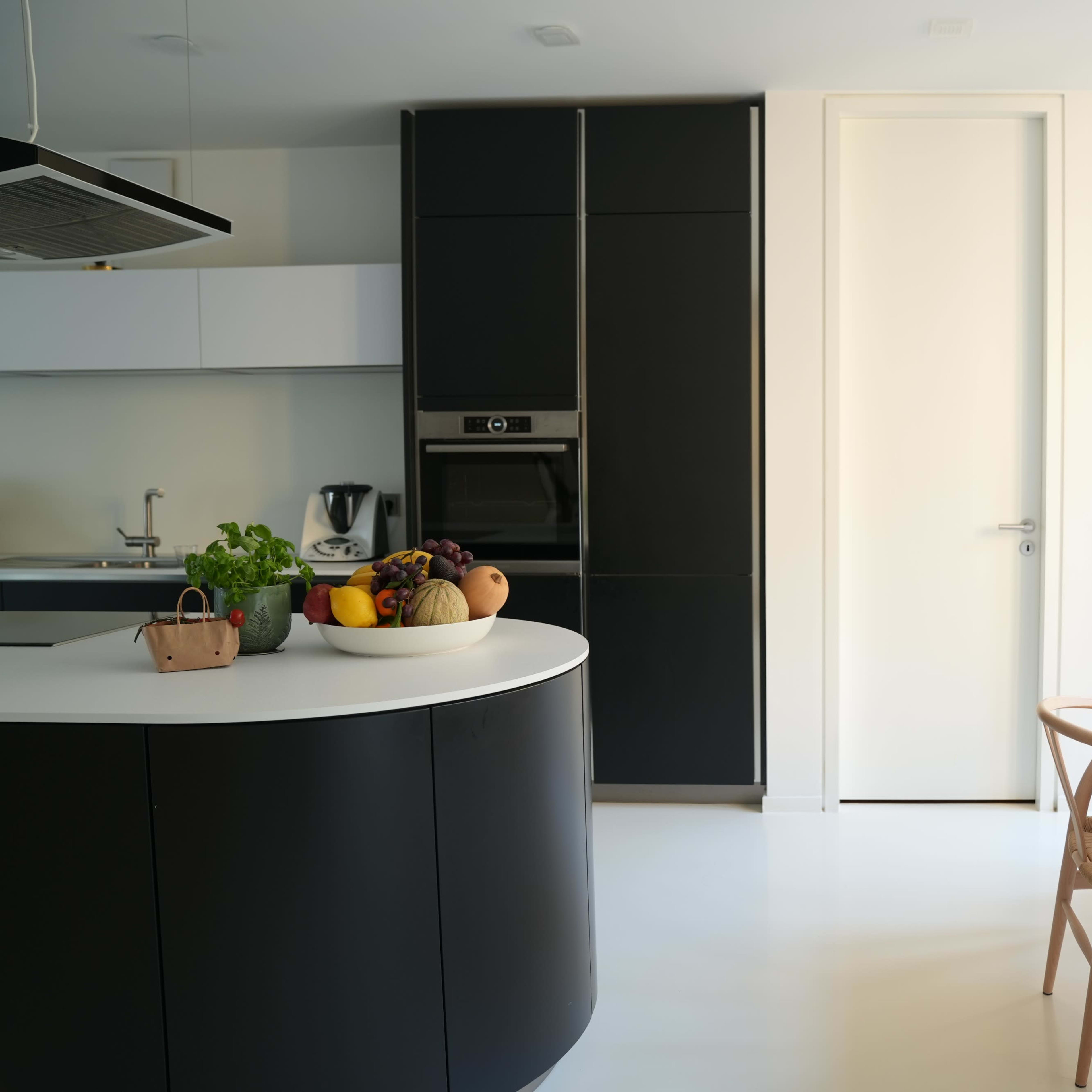 A modern kitchen features a curved island with fruit, sleek cabinetry in black and white, and a minimalist dining table set for a meal.