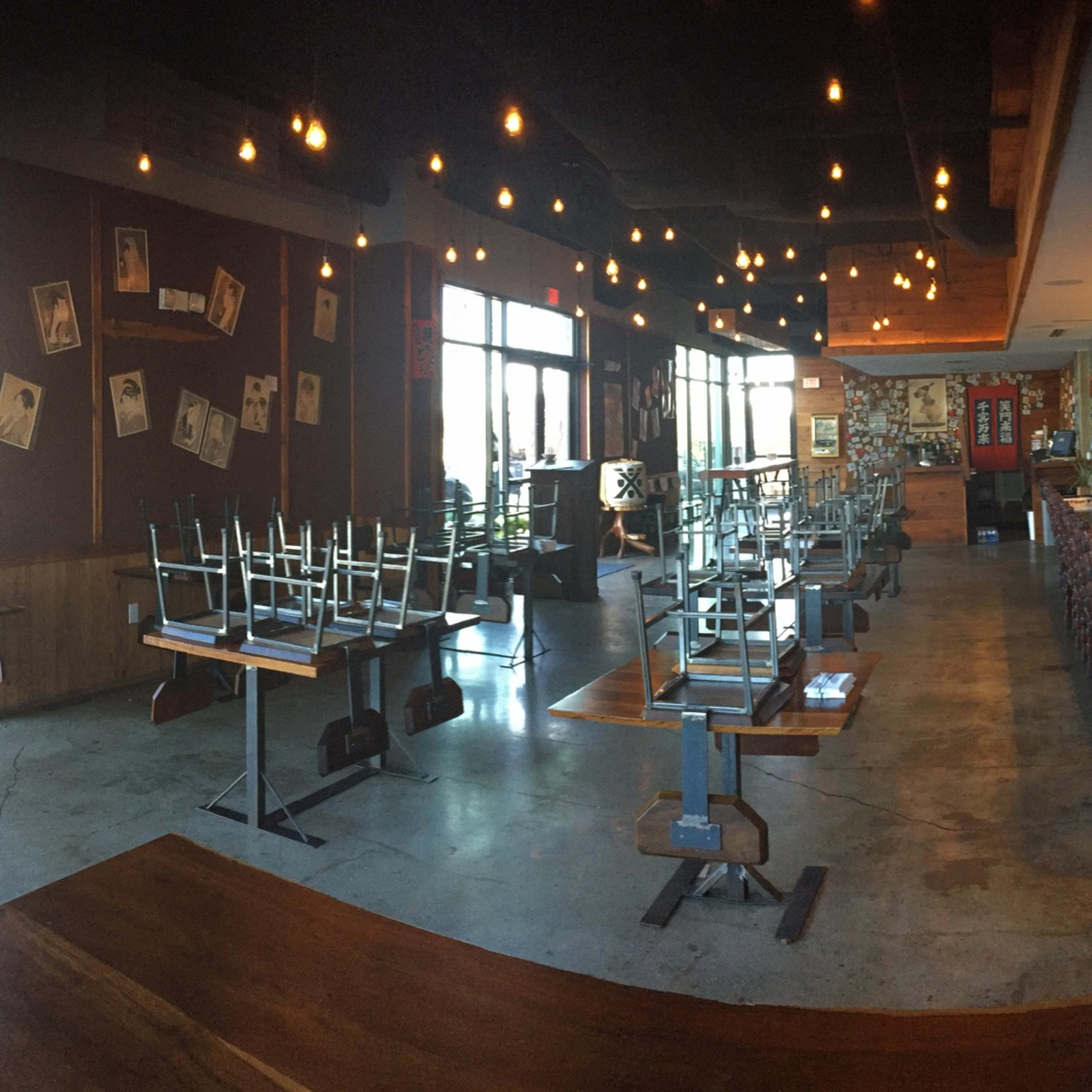 A partially empty restaurant with overturned chairs stacked on tables, exposed light bulbs hanging from the ceiling, and a visible kitchen area at the back.