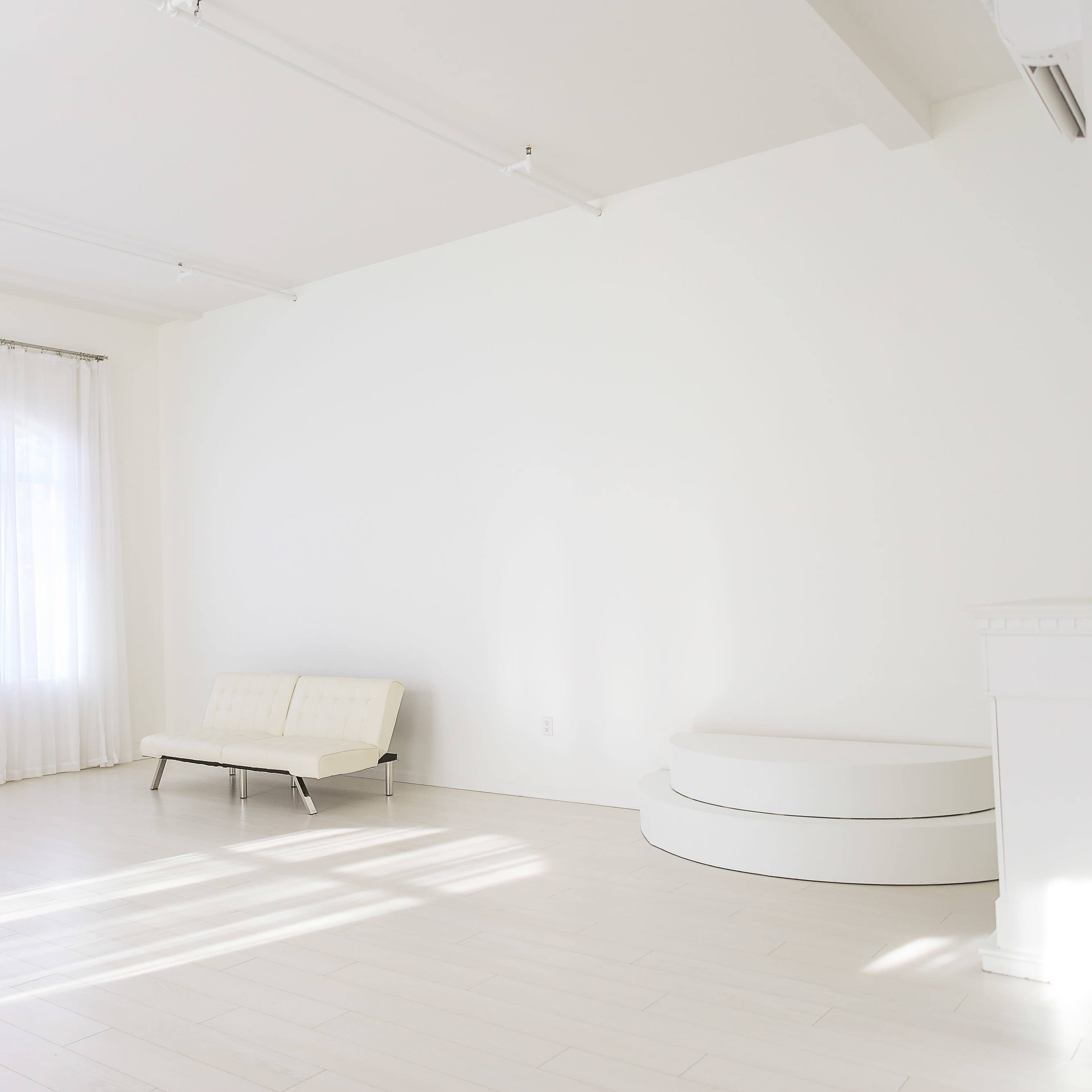 A bright, minimalistic room with light wooden flooring, a white sofa, a circular platform, and large windows covered by sheer curtains.