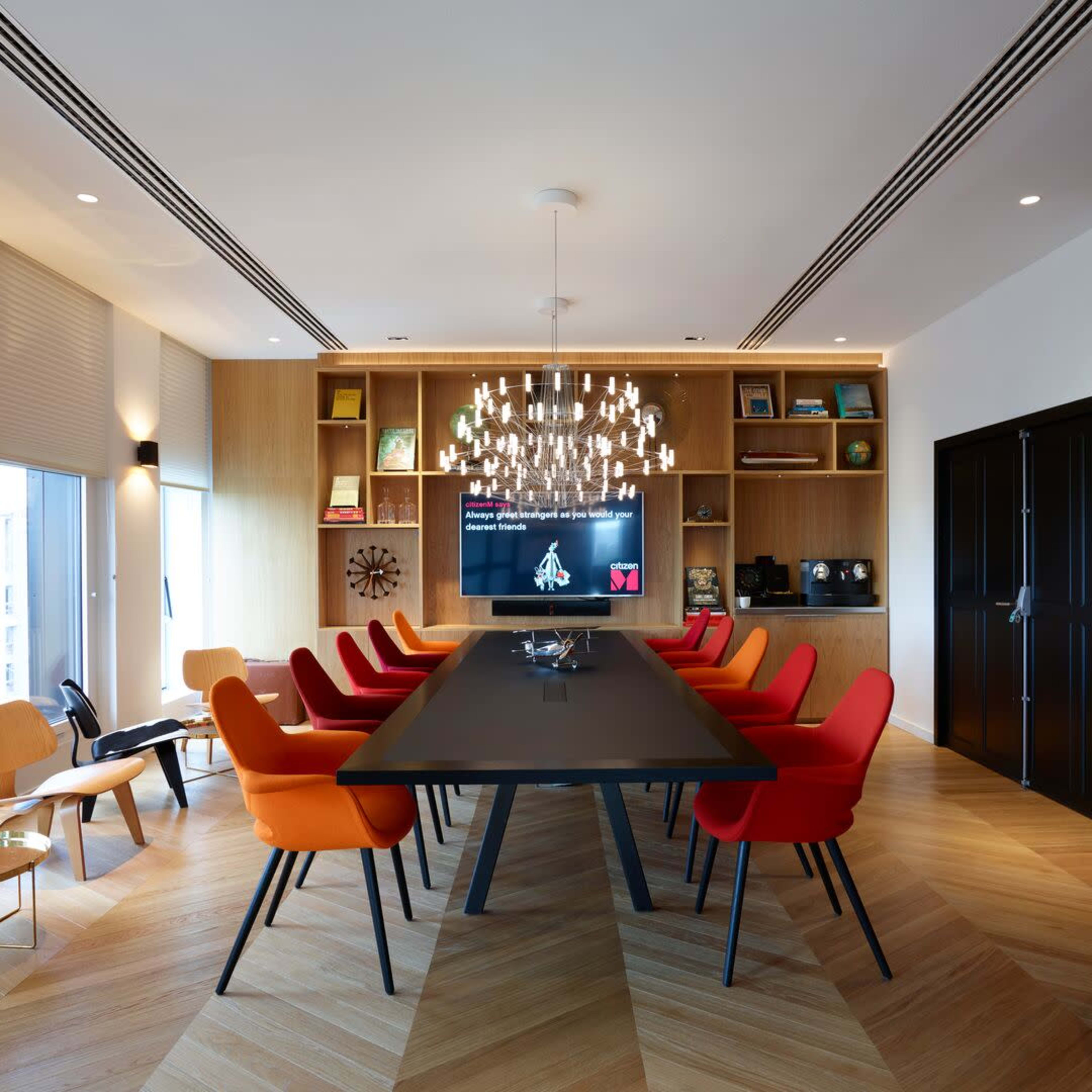 A modern conference room features a large black table surrounded by orange and red chairs, a large screen on the wall, and a stylish chandelier.