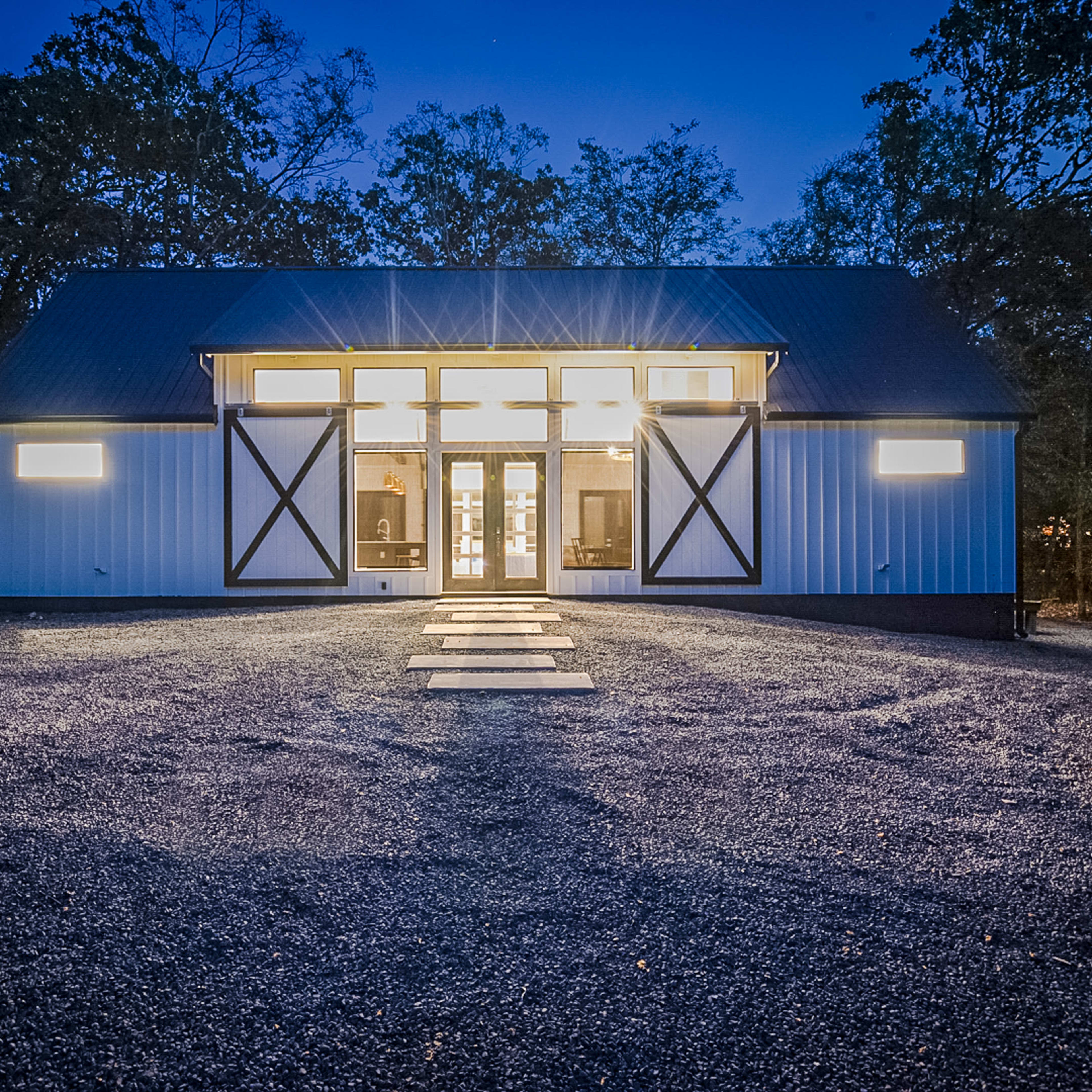 A modern barn-style building is illuminated by large windows at dusk, with a gravel pathway leading to its entrance.