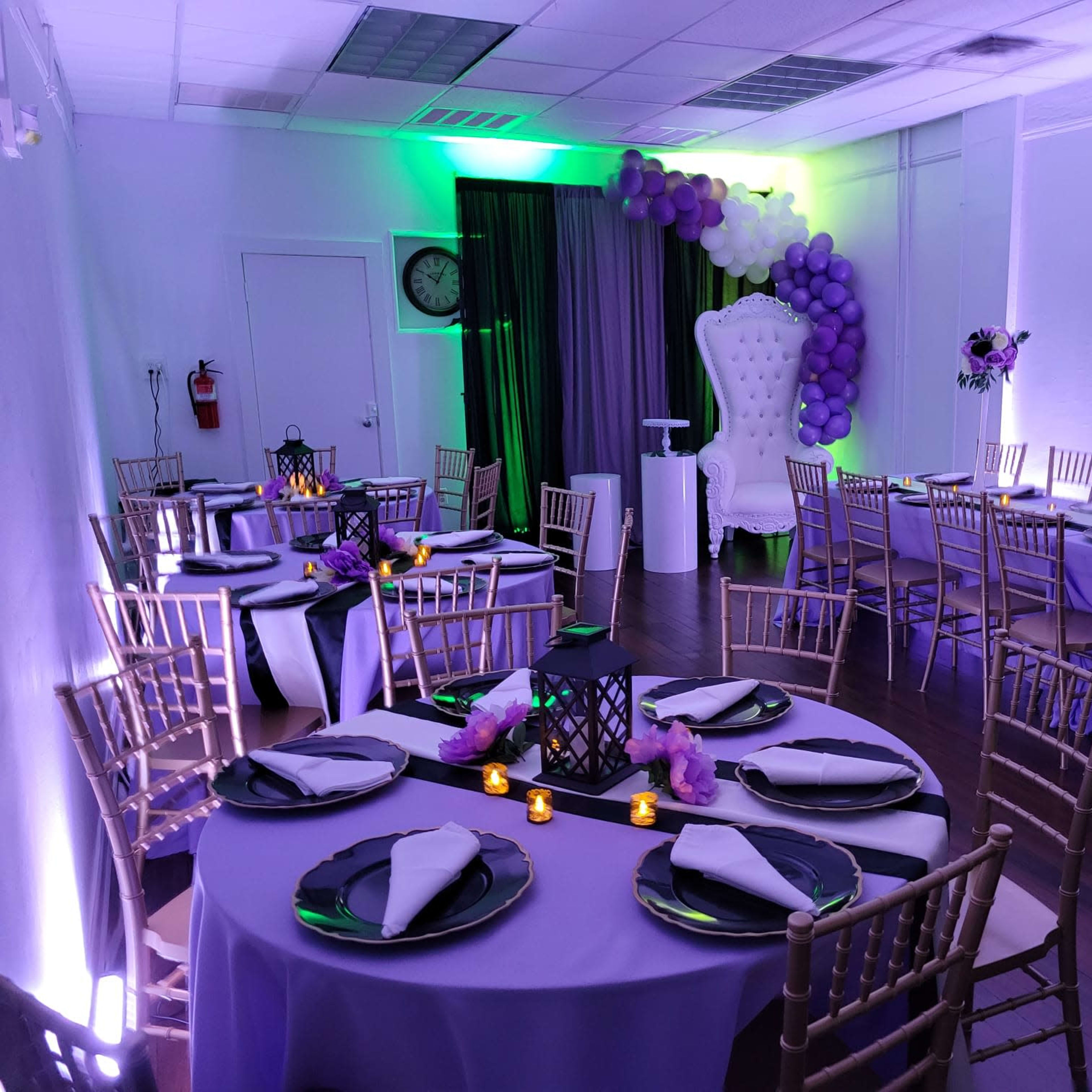 A banquet hall is elegantly decorated with purple and white balloons, tables set with black plates and napkins, and a large white chair at the front.