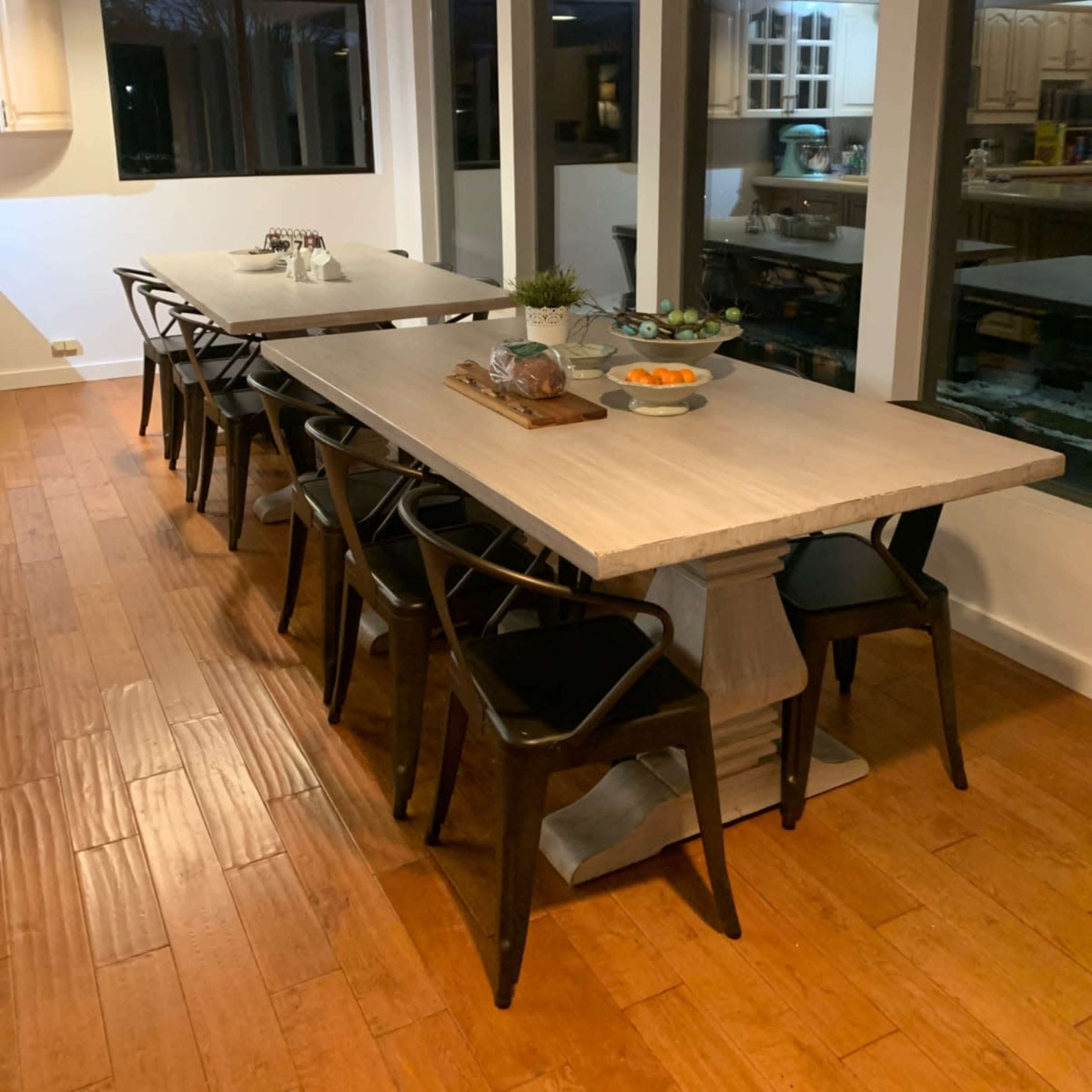 A long dining table with black chairs is set against a backdrop of large windows in a modern kitchen.
