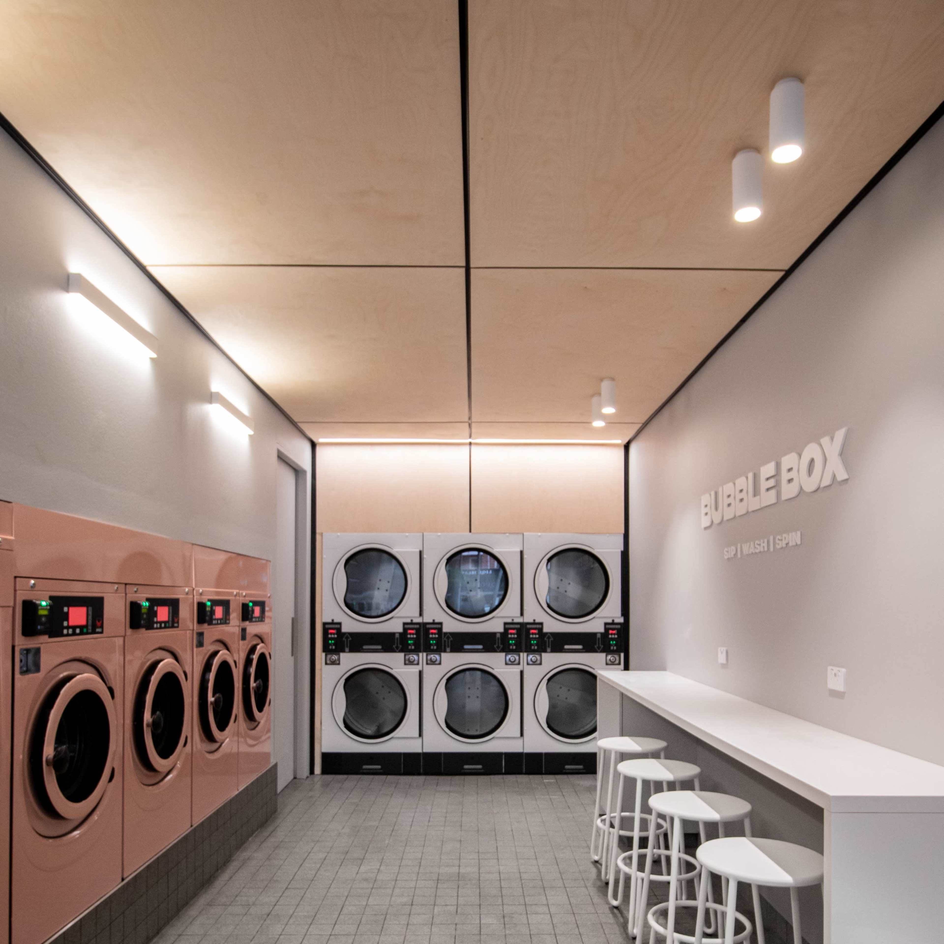 The interior of a modern laundromat features rows of washing machines and dryers against a minimalist wall, with stools and a counter for customers.