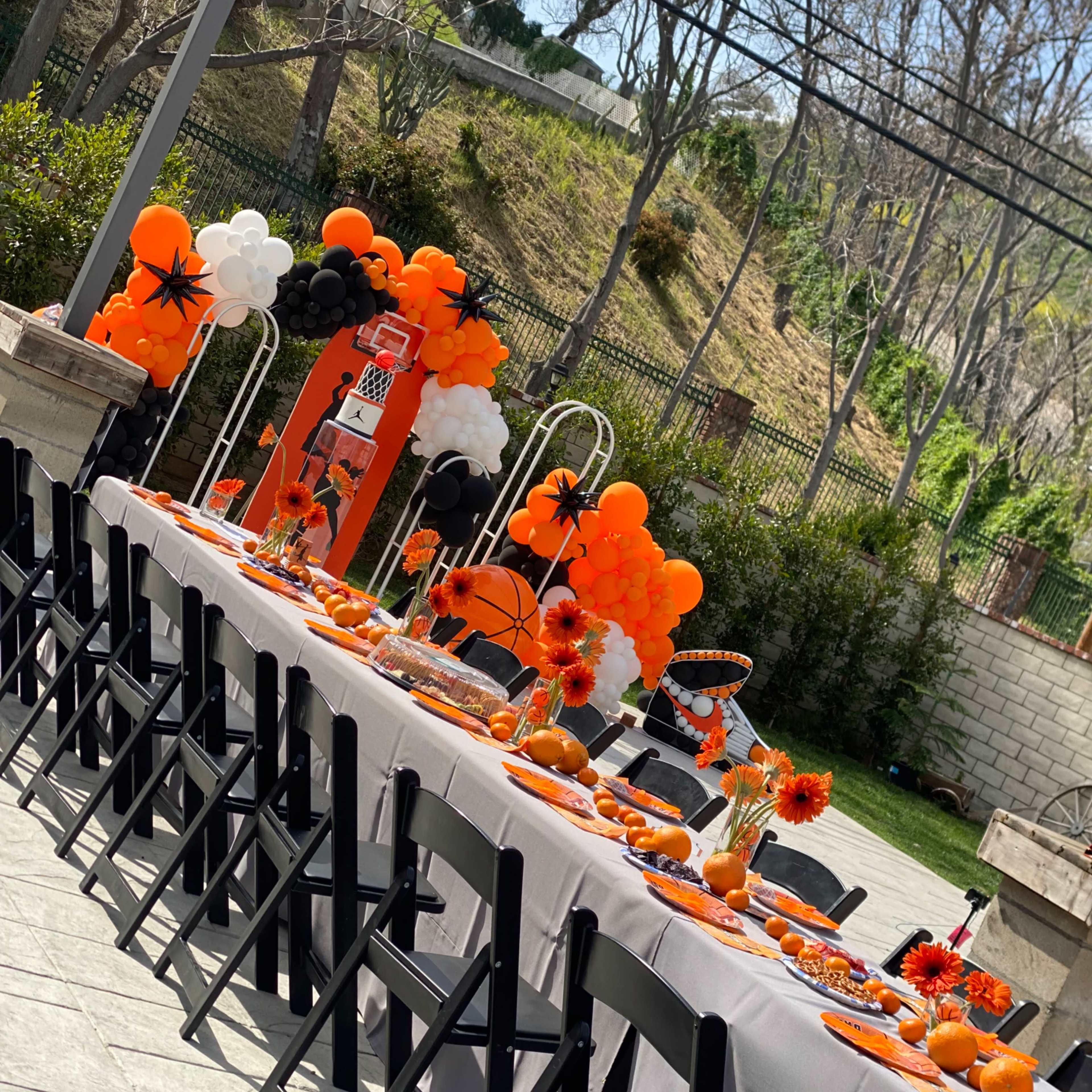 A long outdoor table is set up with an orange and black themed decoration, featuring flowers, balloons, and various party items.