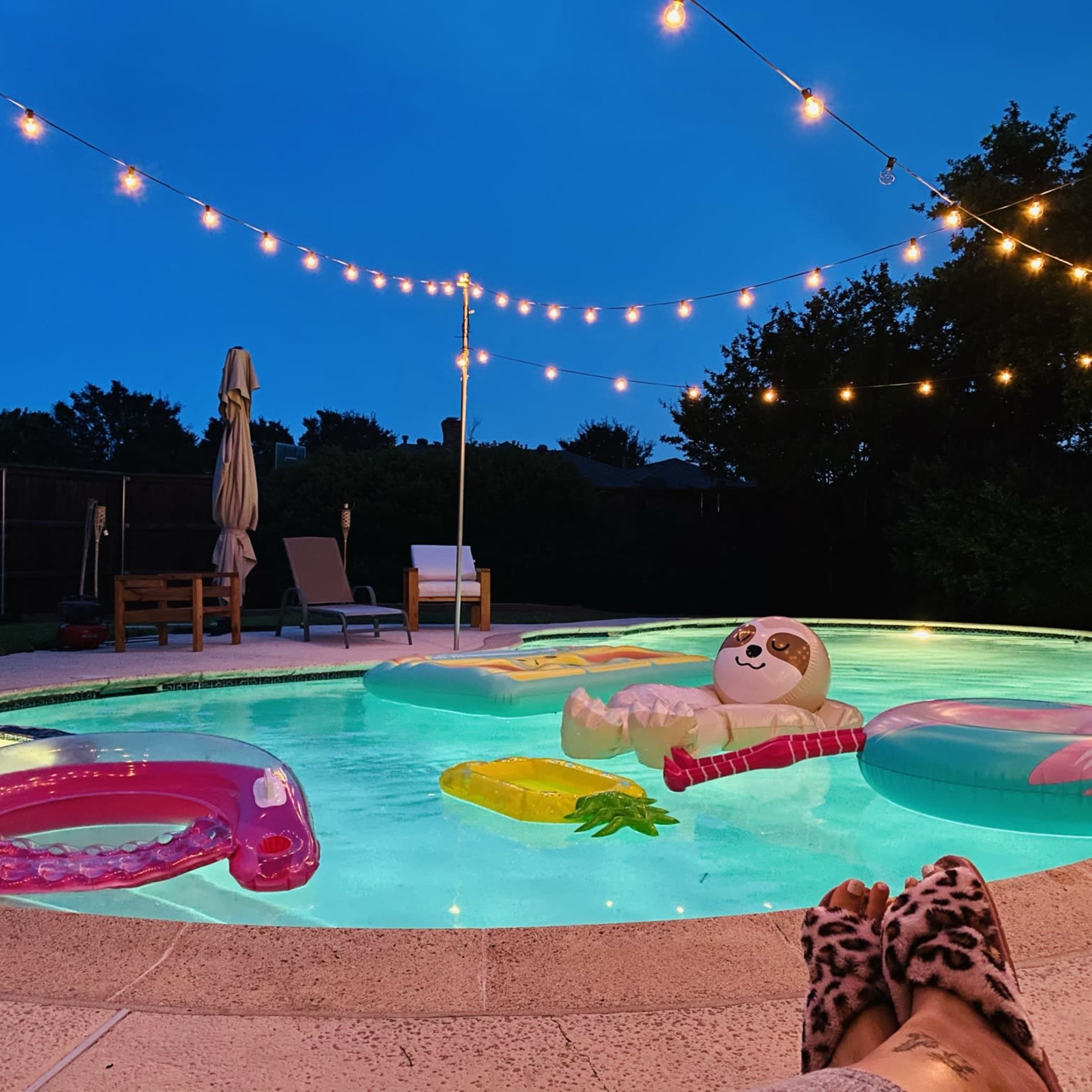 The scene shows a brightly lit pool at dusk, adorned with floating toys and string lights overhead.