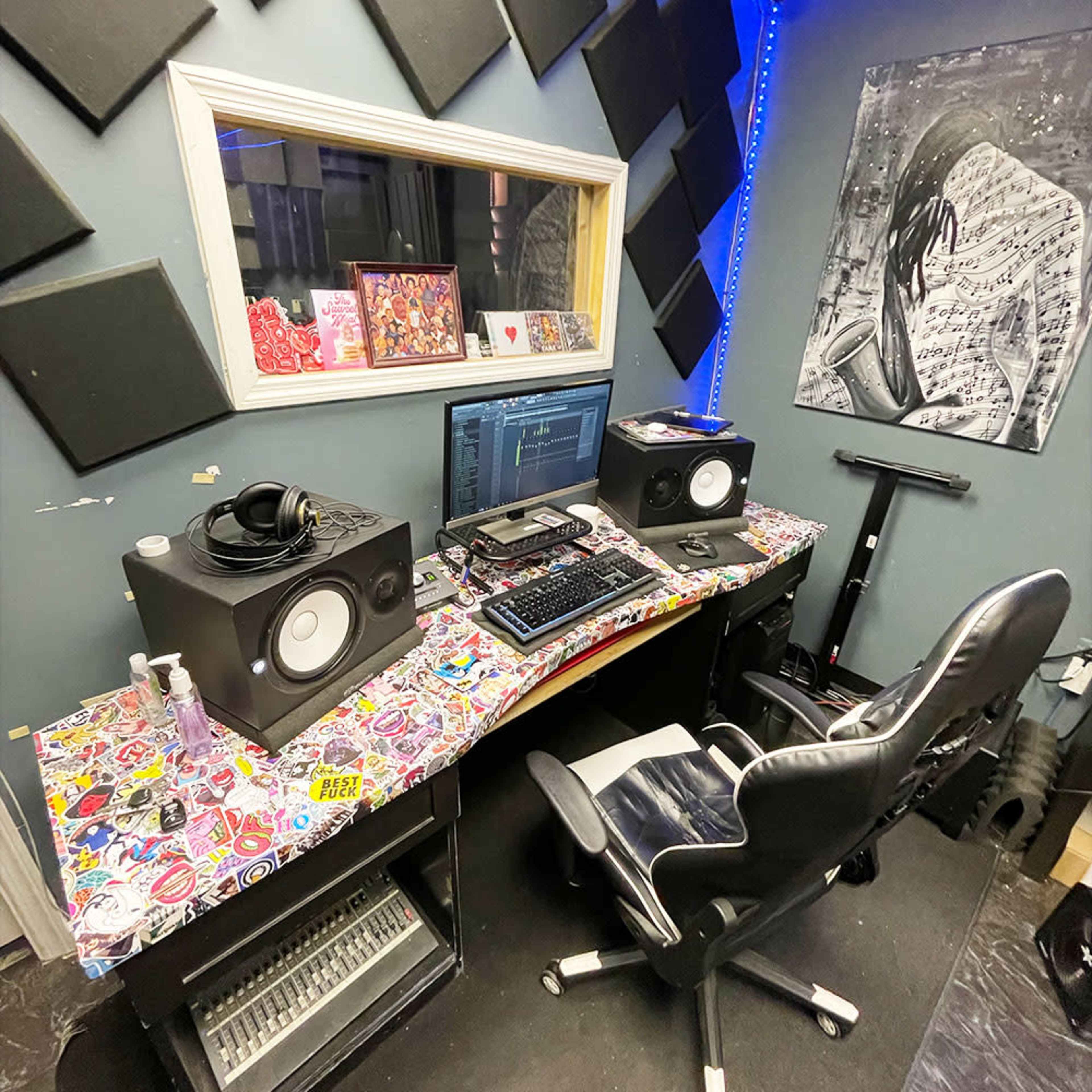 A music production studio with a desk featuring two speakers, a computer monitor, a keyboard, a decorated tabletop covered in stickers, and a black office chair.