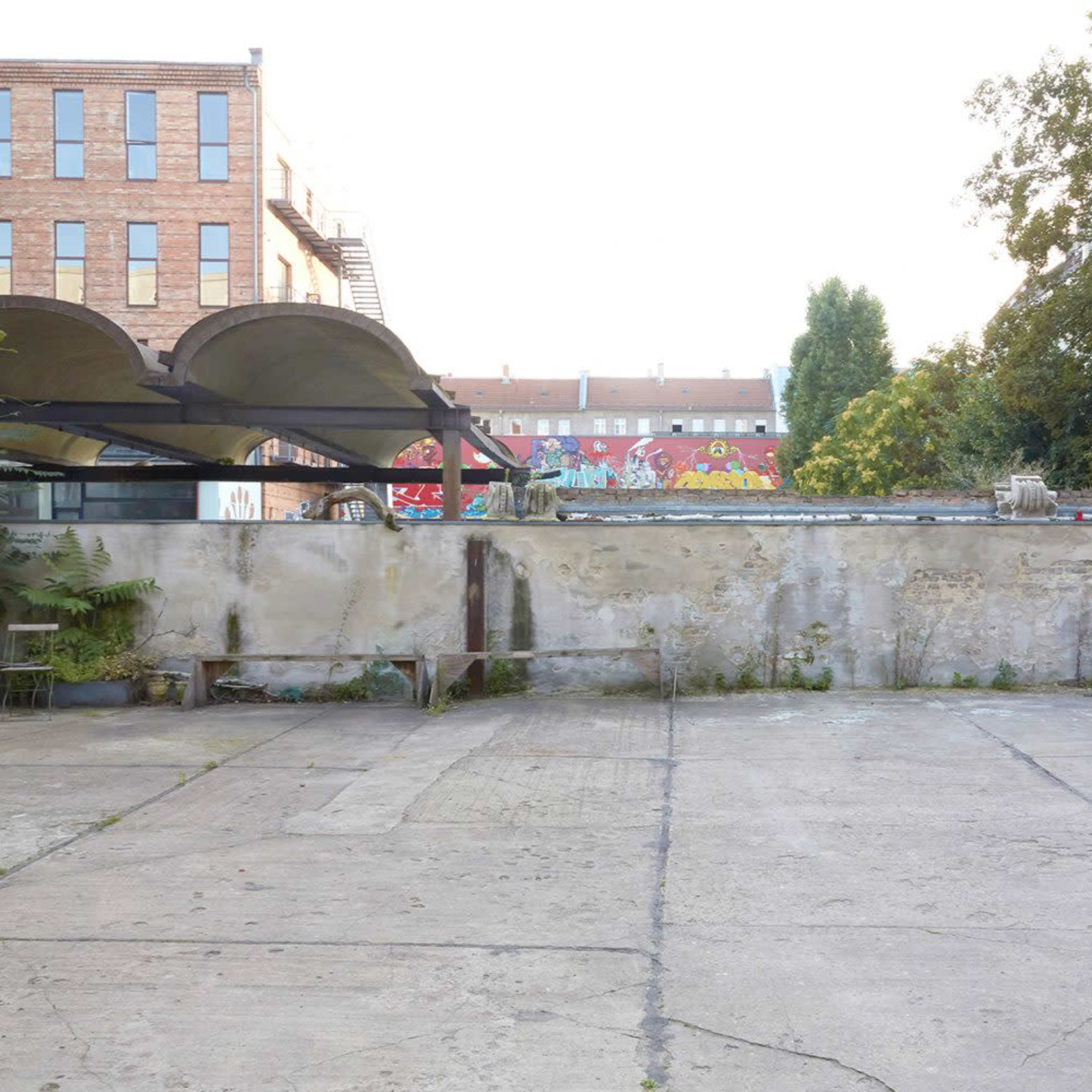 An empty outdoor space with a concrete floor and a weathered wall, partially covered by greenery and graffiti in the background.