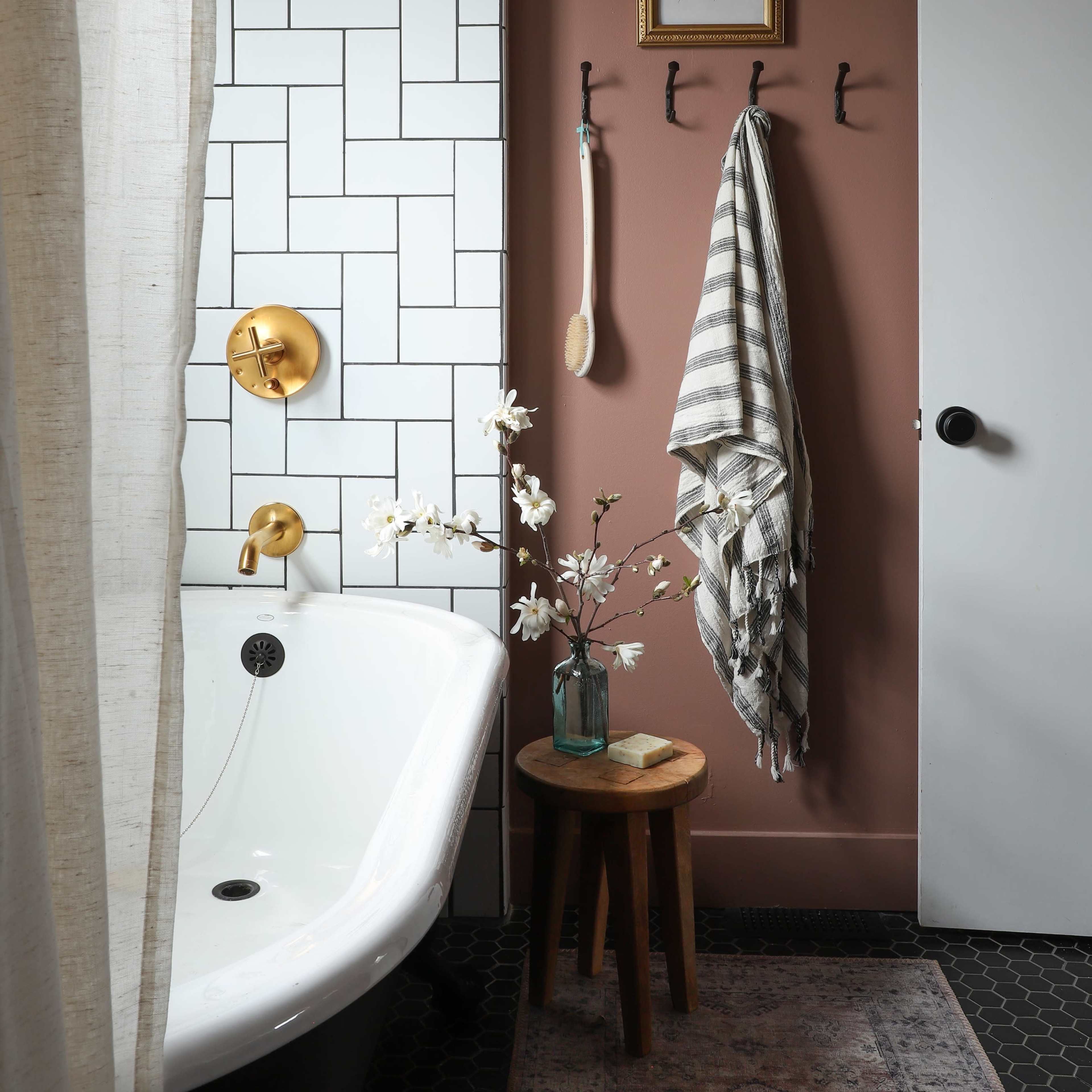 A bathroom featuring a freestanding bathtub, decorative wall tiles, a dark rug, and a wooden stool with a vase of flowers.