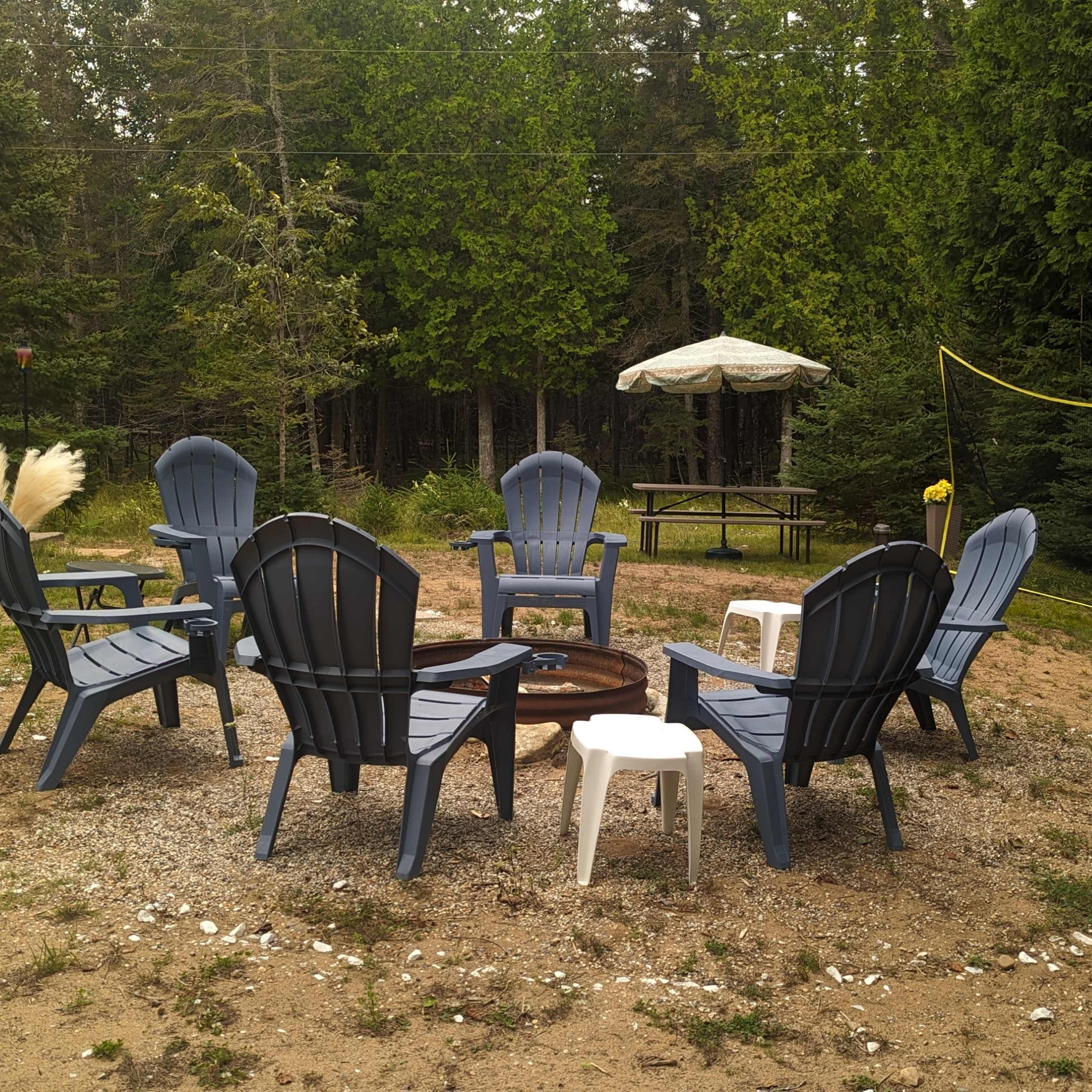A circle of eight blue Adirondack chairs surrounds a fire pit in a clearing surrounded by trees, with a picnic table and an umbrella in the background.