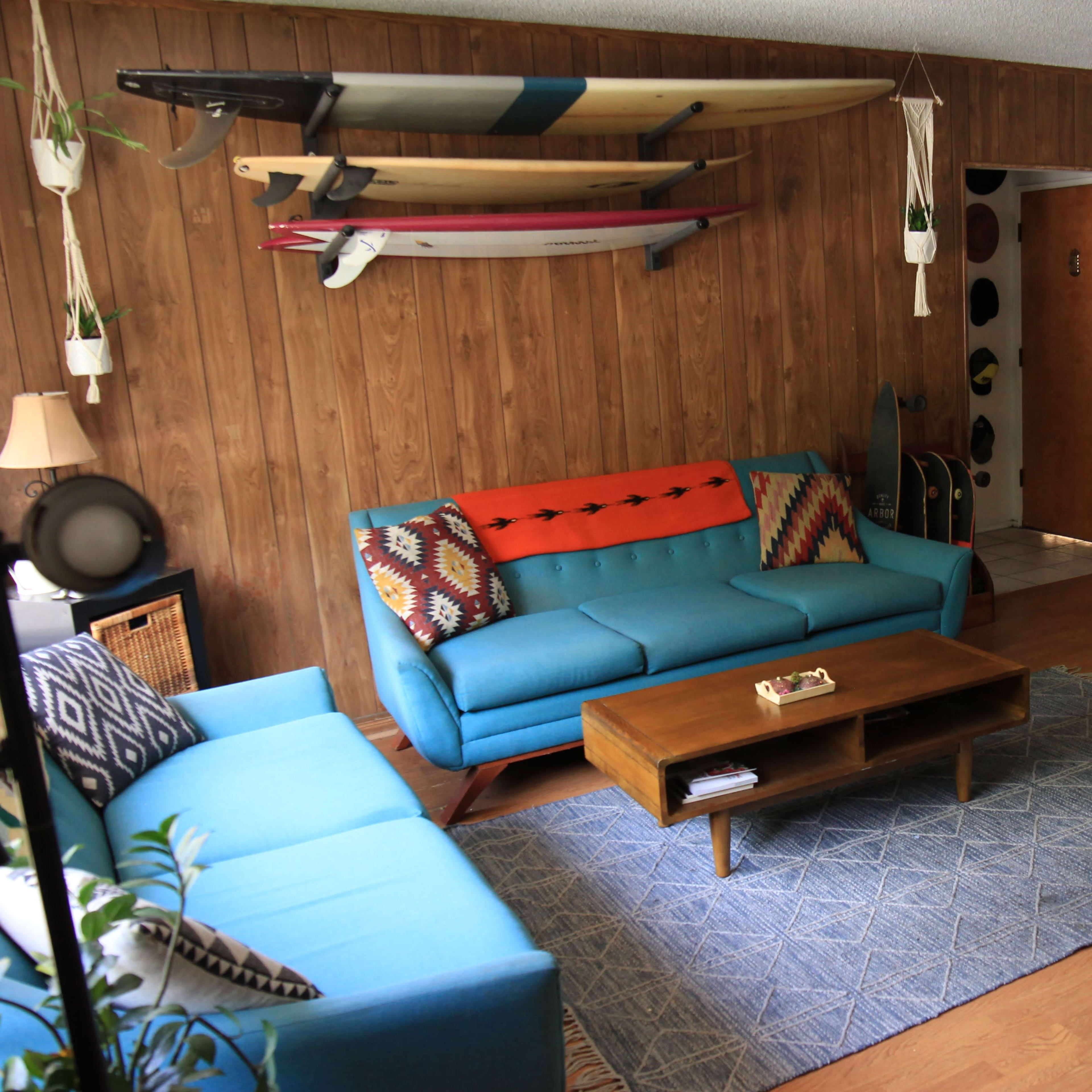 A living room features a blue sofa, a wooden coffee table, and surfboards mounted on the wall, with a vintage piano and plants enhancing the space.