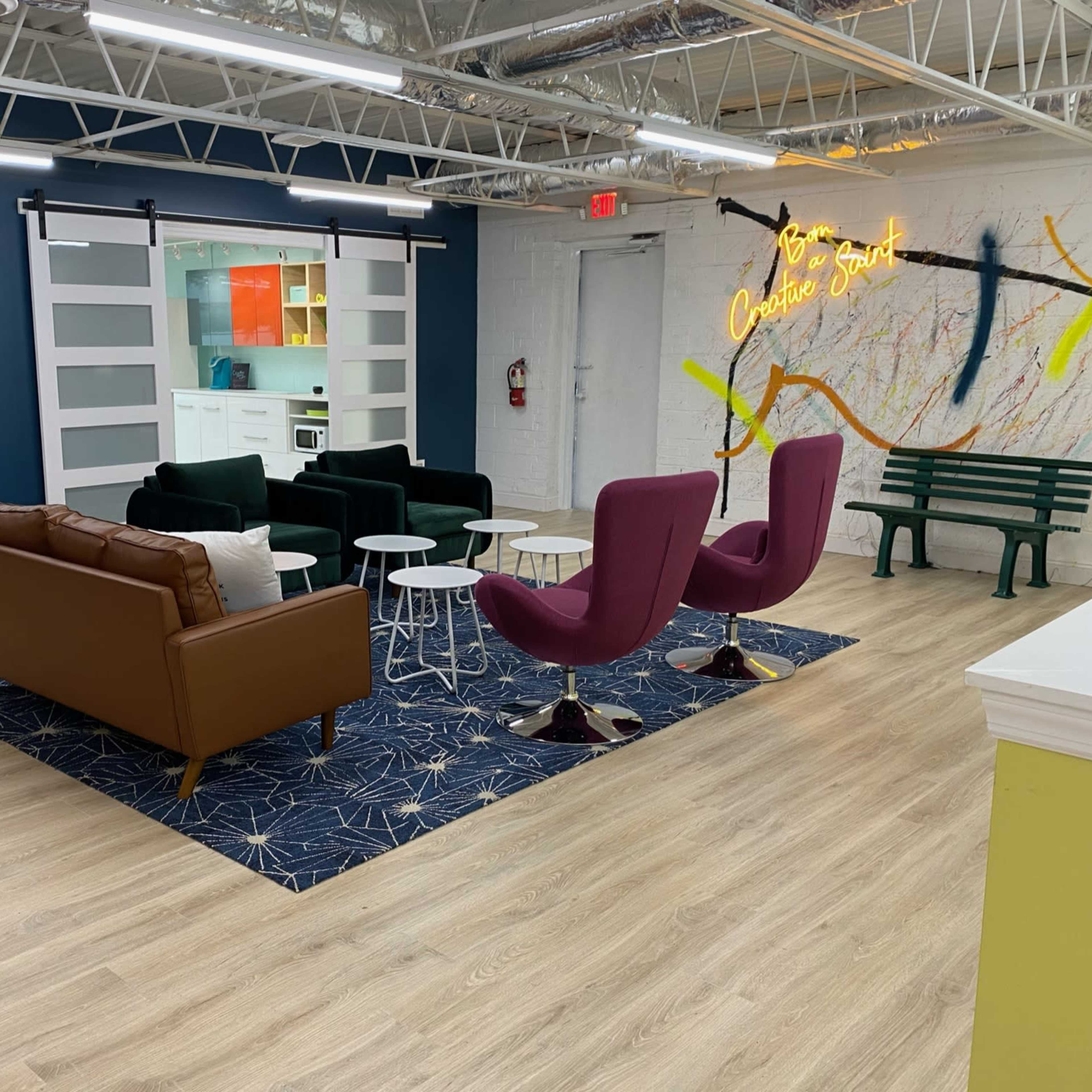 A modern lounge area with a mix of seating options, including a brown leather couch, purple armchairs, and a green bench, situated on a patterned rug in a well-lit space.