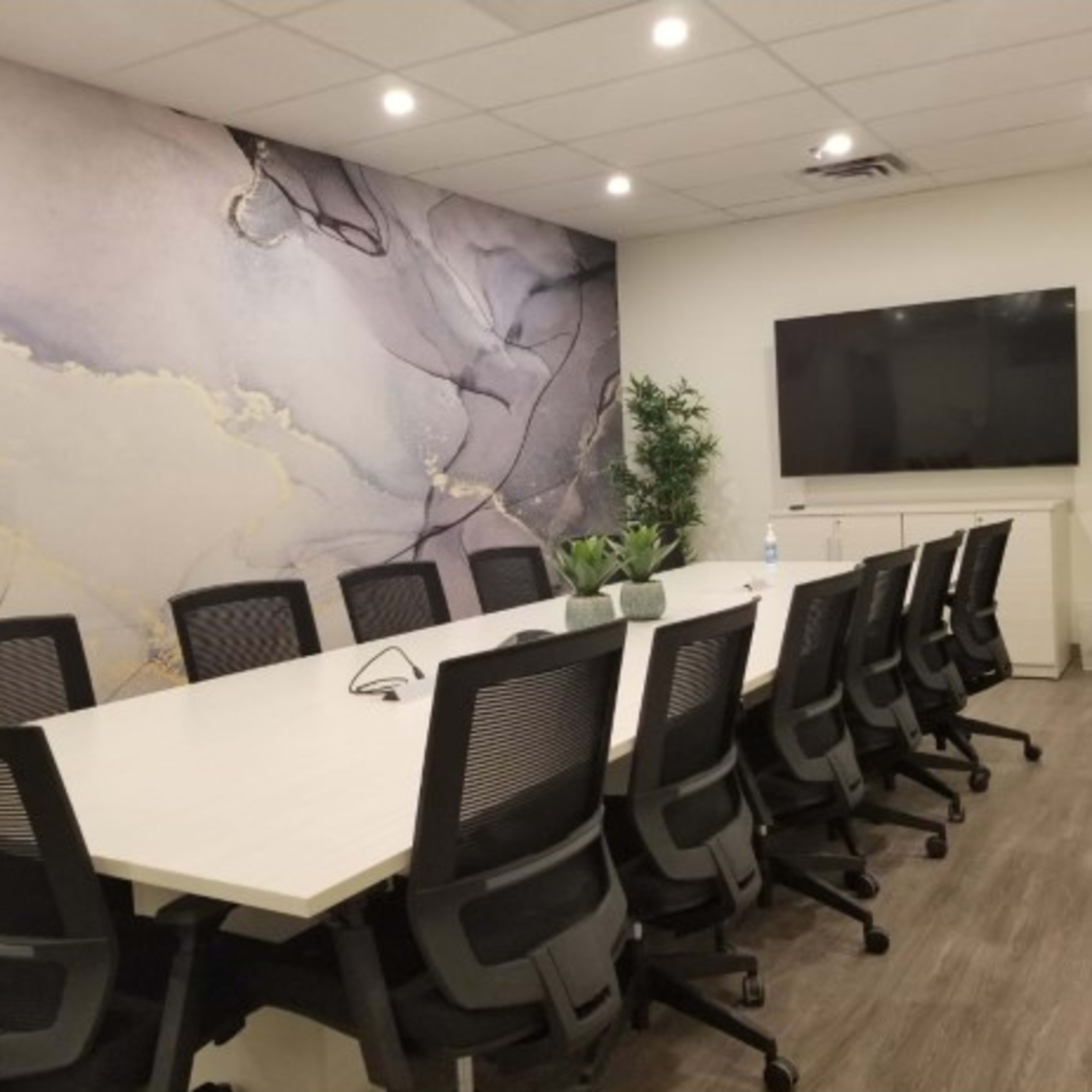 A modern conference room featuring a long white table surrounded by black ergonomic chairs, a large wall-mounted screen, and a decorative wall with abstract patterns.