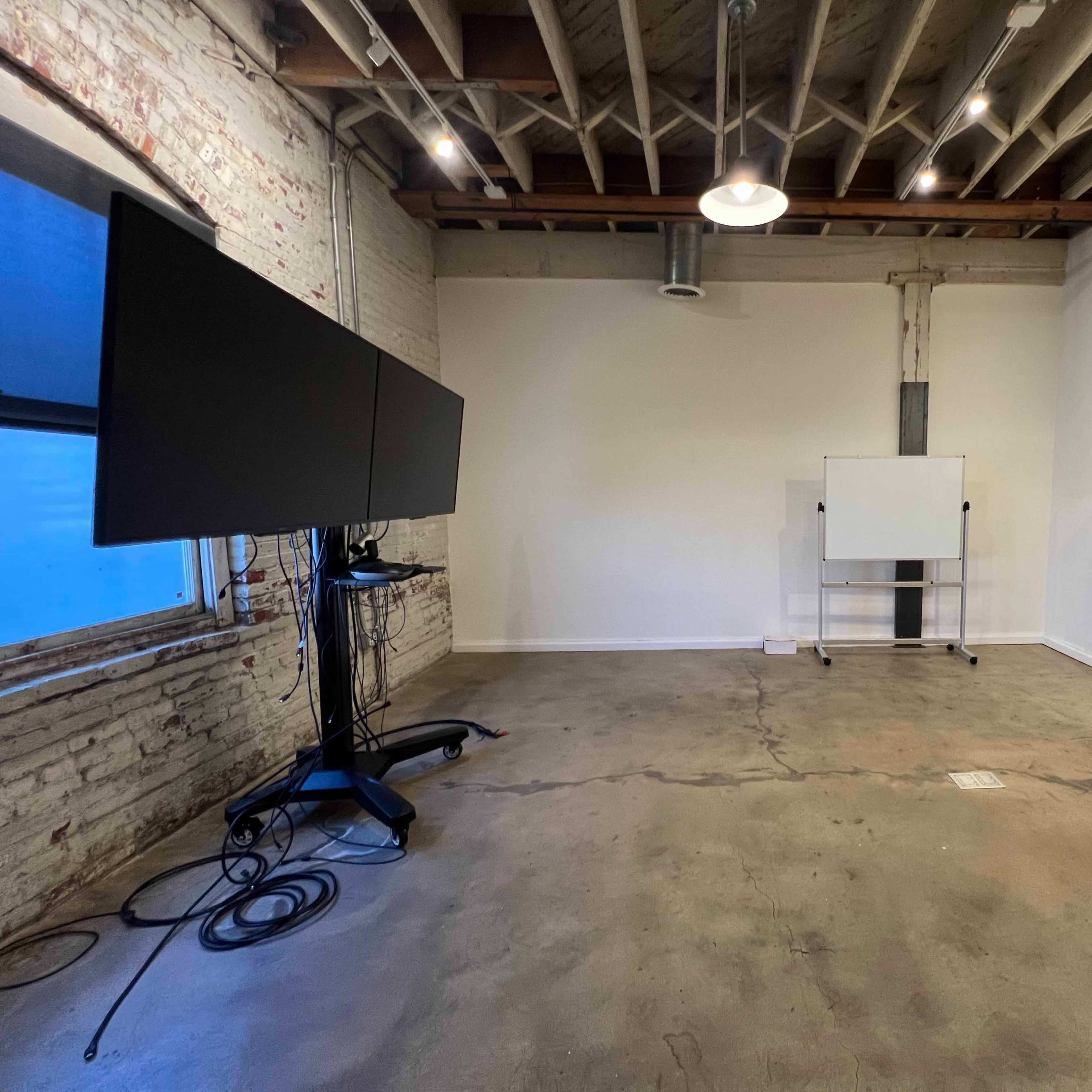 An empty office space features a large monitor on a stand, a whiteboard on wheels, and a wooden shelving unit against the wall.