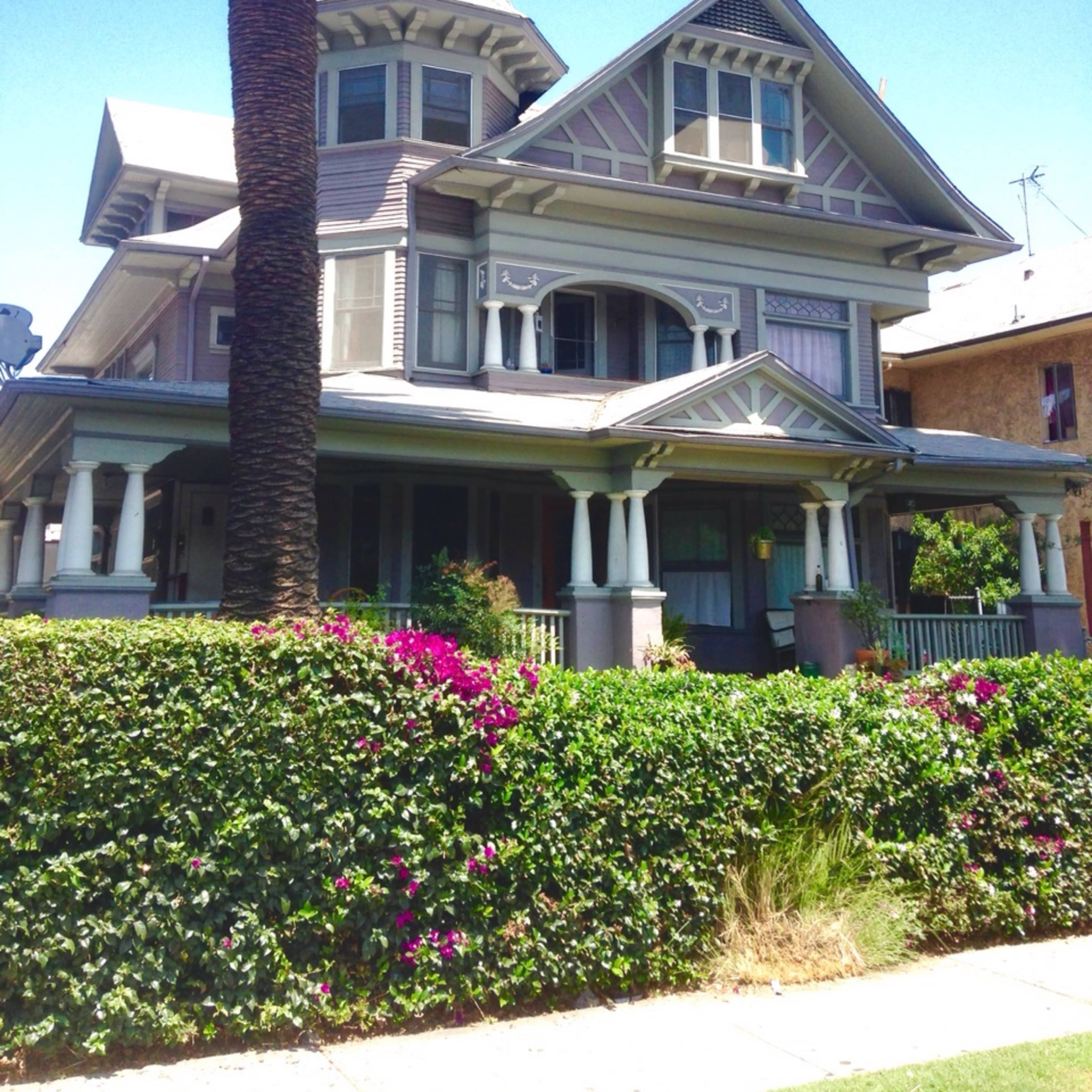 A large, multi-story Victorian-style house with a turret, surrounded by green shrubs and flowering plants, under a clear blue sky.
