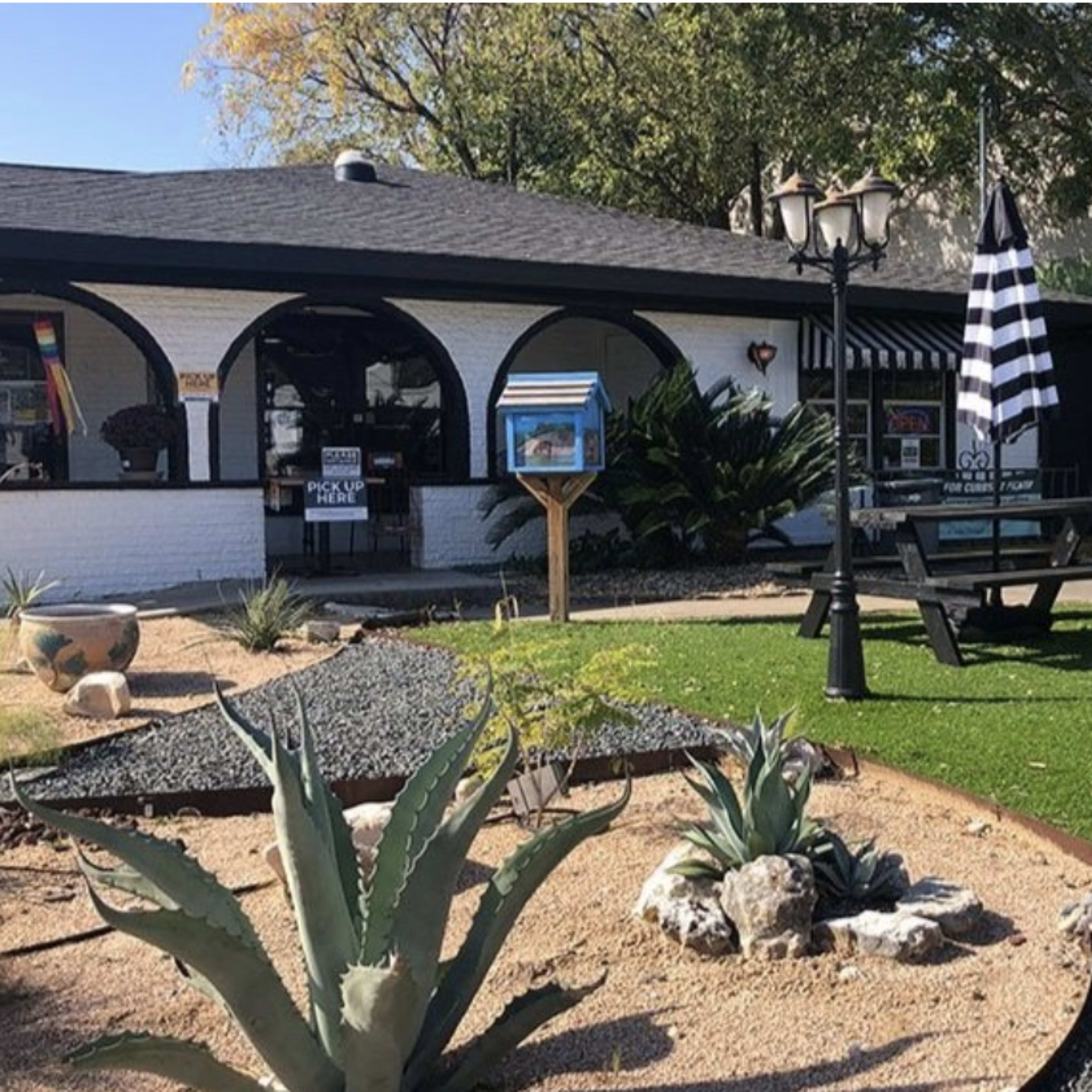 A landscaped outdoor area with black and white striped umbrellas over picnic tables, bordered by gravel and plants in front of a white building.