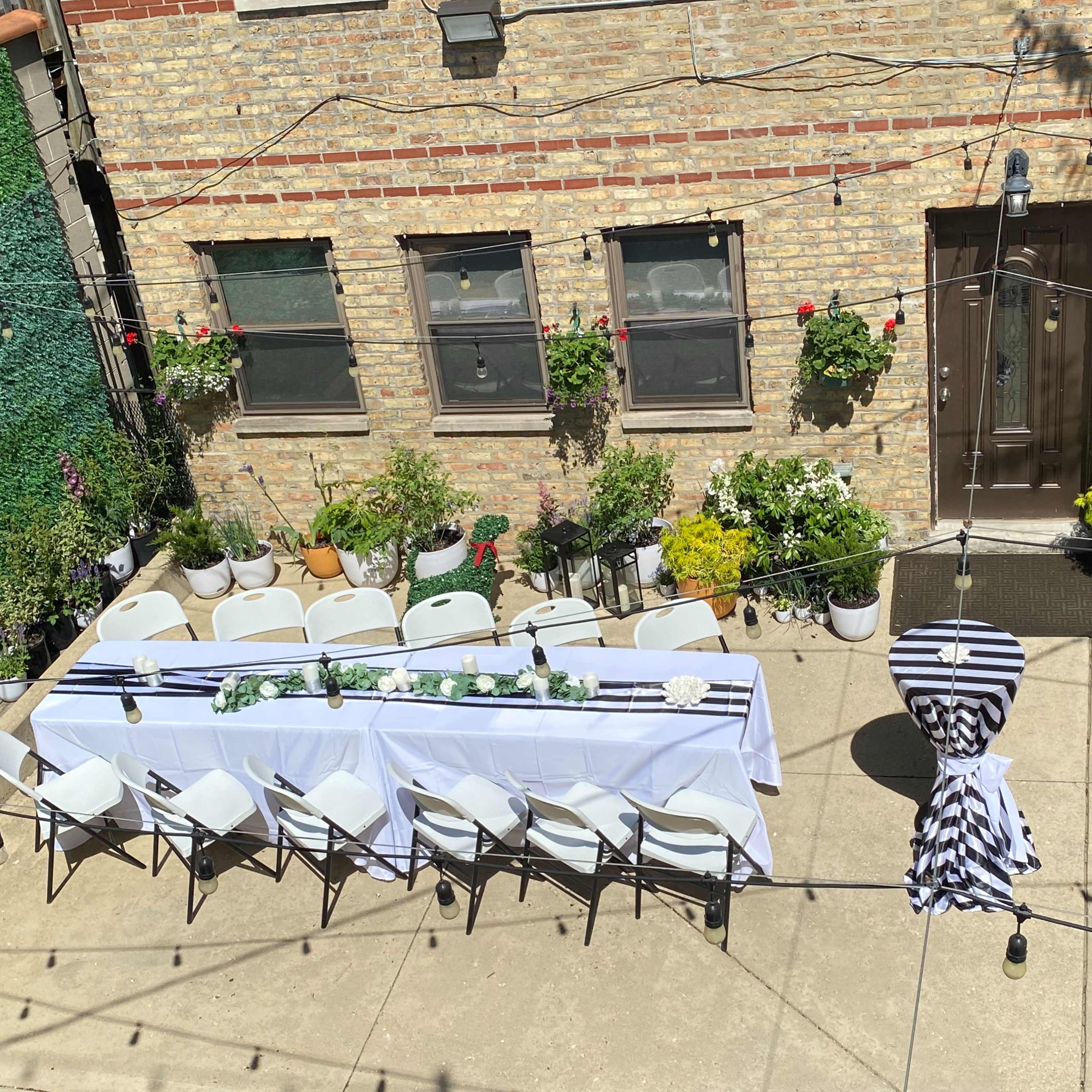 A long table set with white linens and chairs is arranged in a garden patio surrounded by potted plants and hanging flowers.