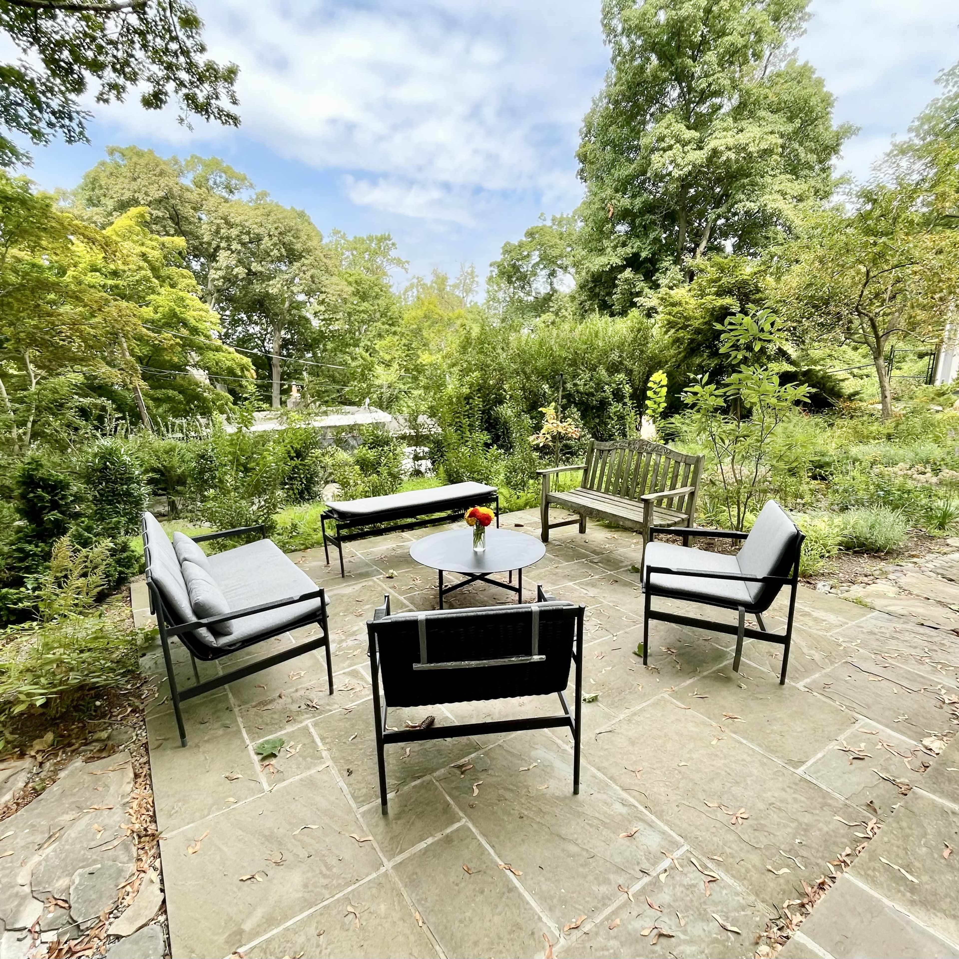 A stone patio features several black outdoor chairs and a table, surrounded by greenery and trees.