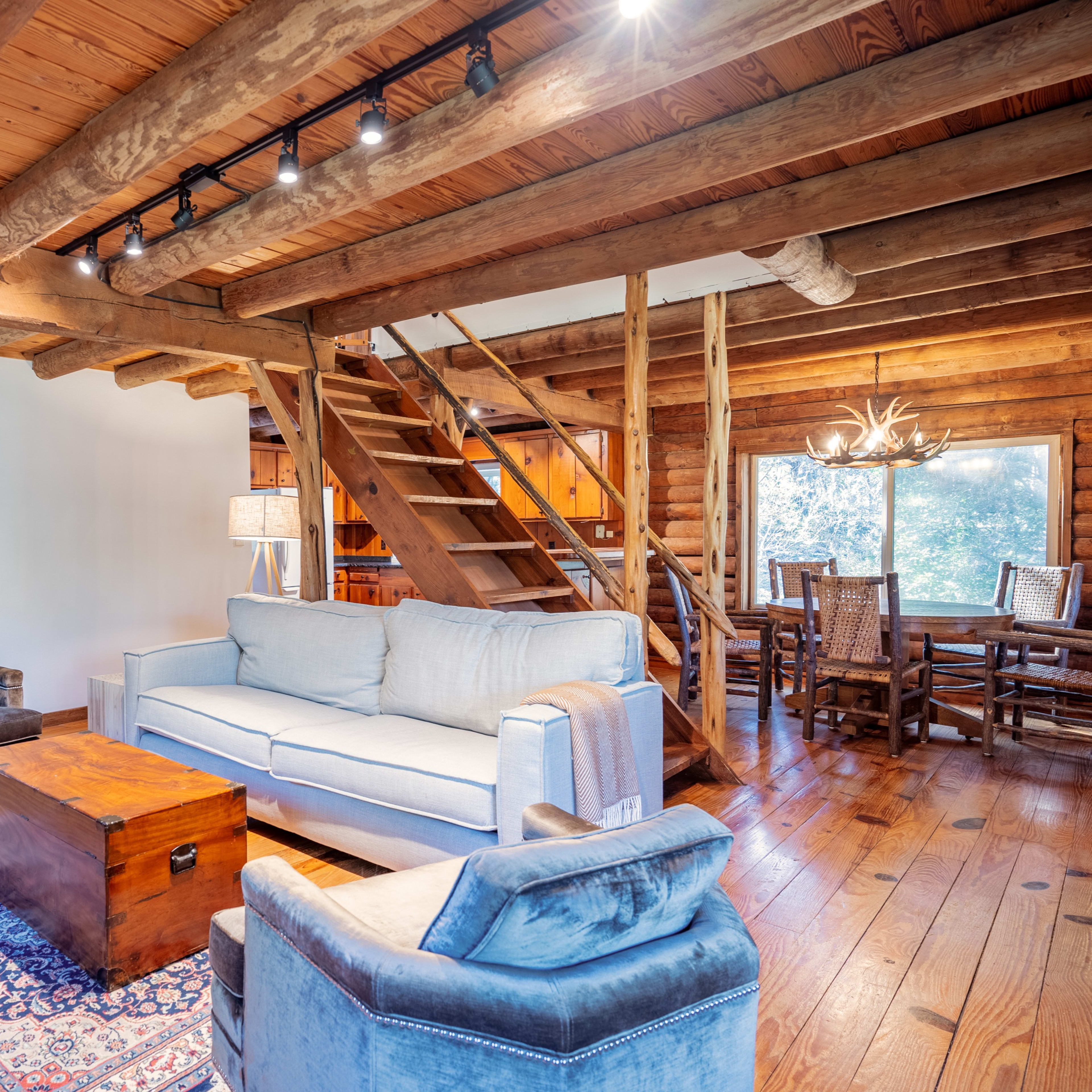 A rustic living room with wooden beams, a light-colored sofa, a wooden coffee table, and a staircase leading to an upper level.
