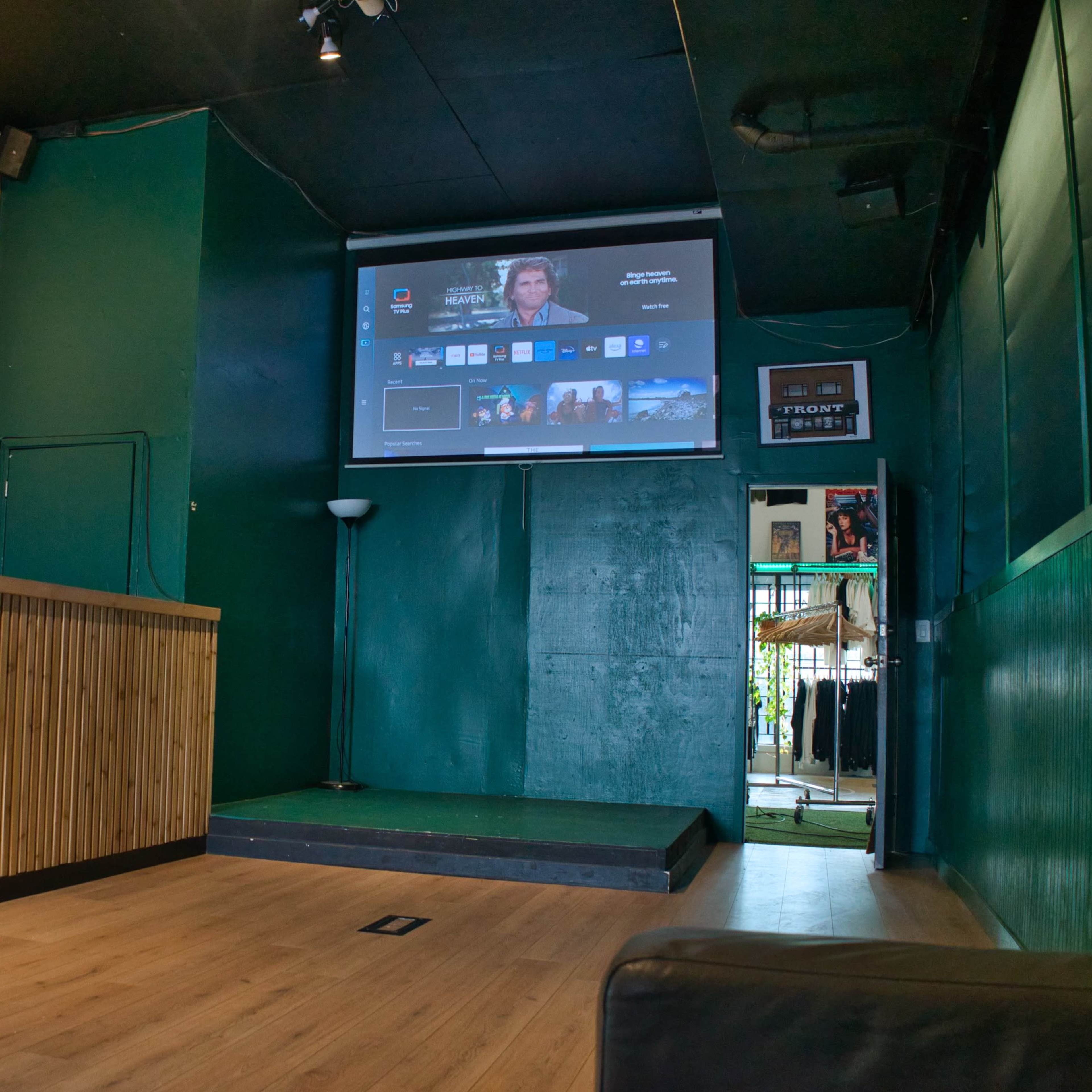 A dimly lit room with a wooden reception desk, a large screen displaying content on a green wall, and a small stage area.
