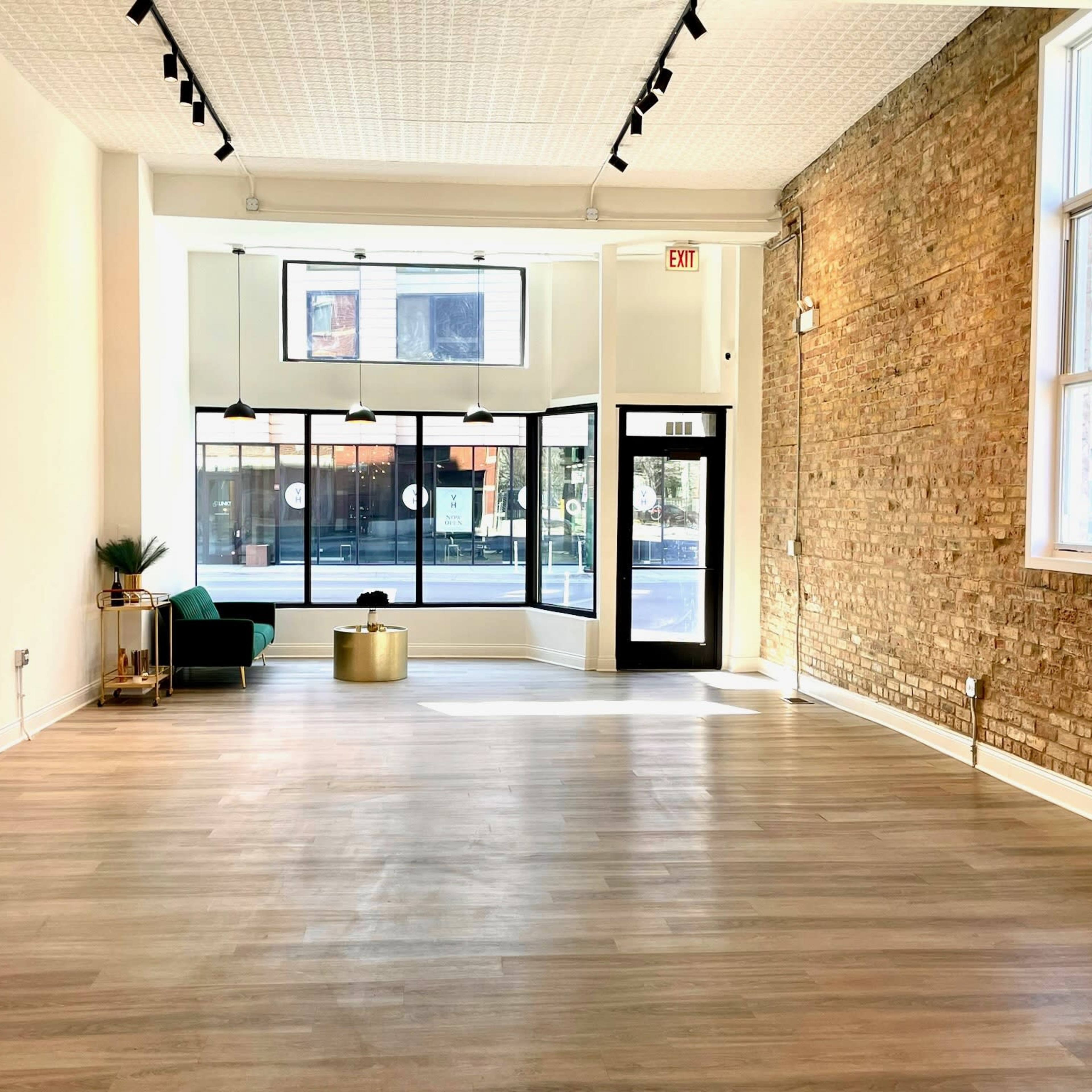 An empty, well-lit interior space with wooden flooring, large front windows, and exposed brick walls.