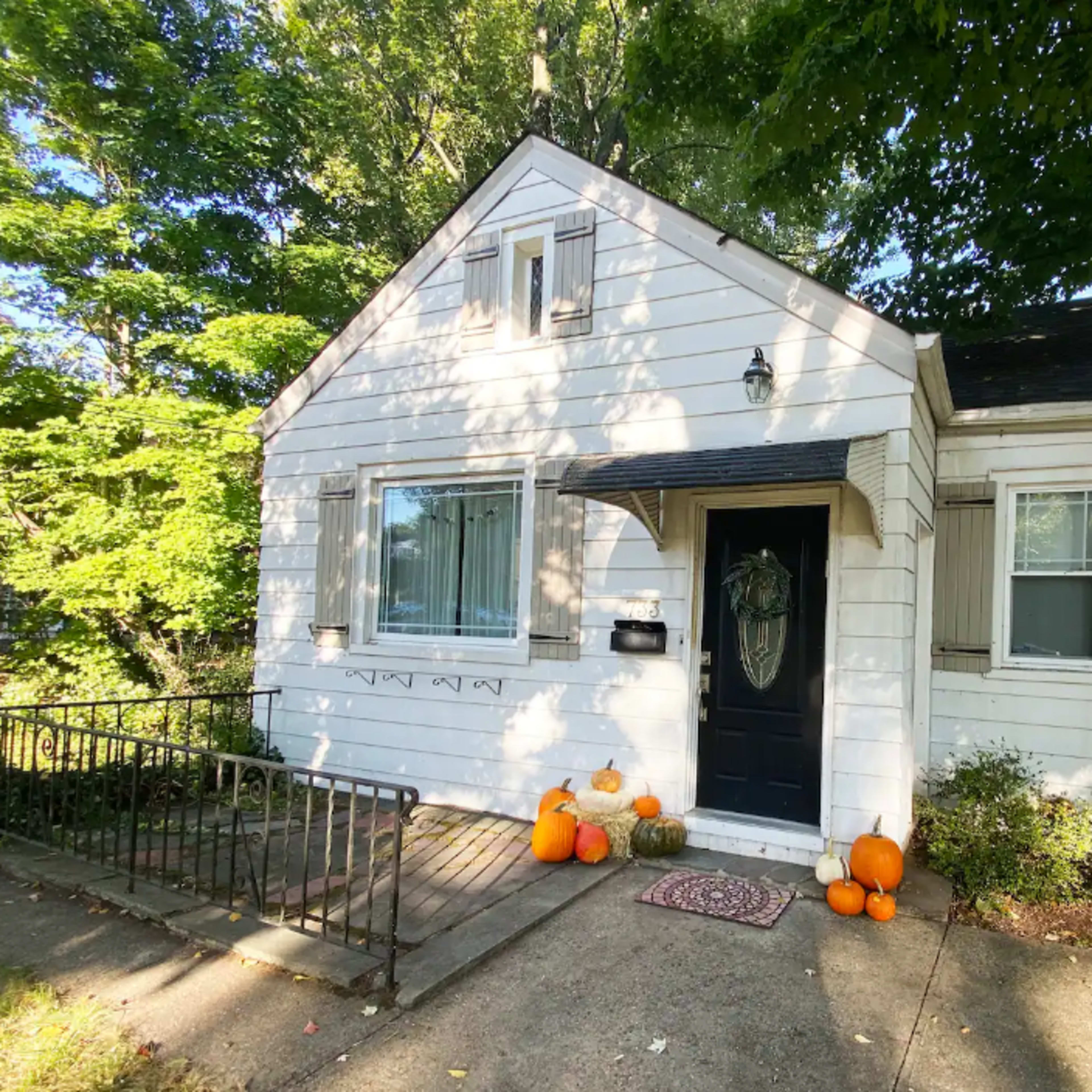 A small white house with a black door has pumpkins arranged on the porch and is surrounded by trees.