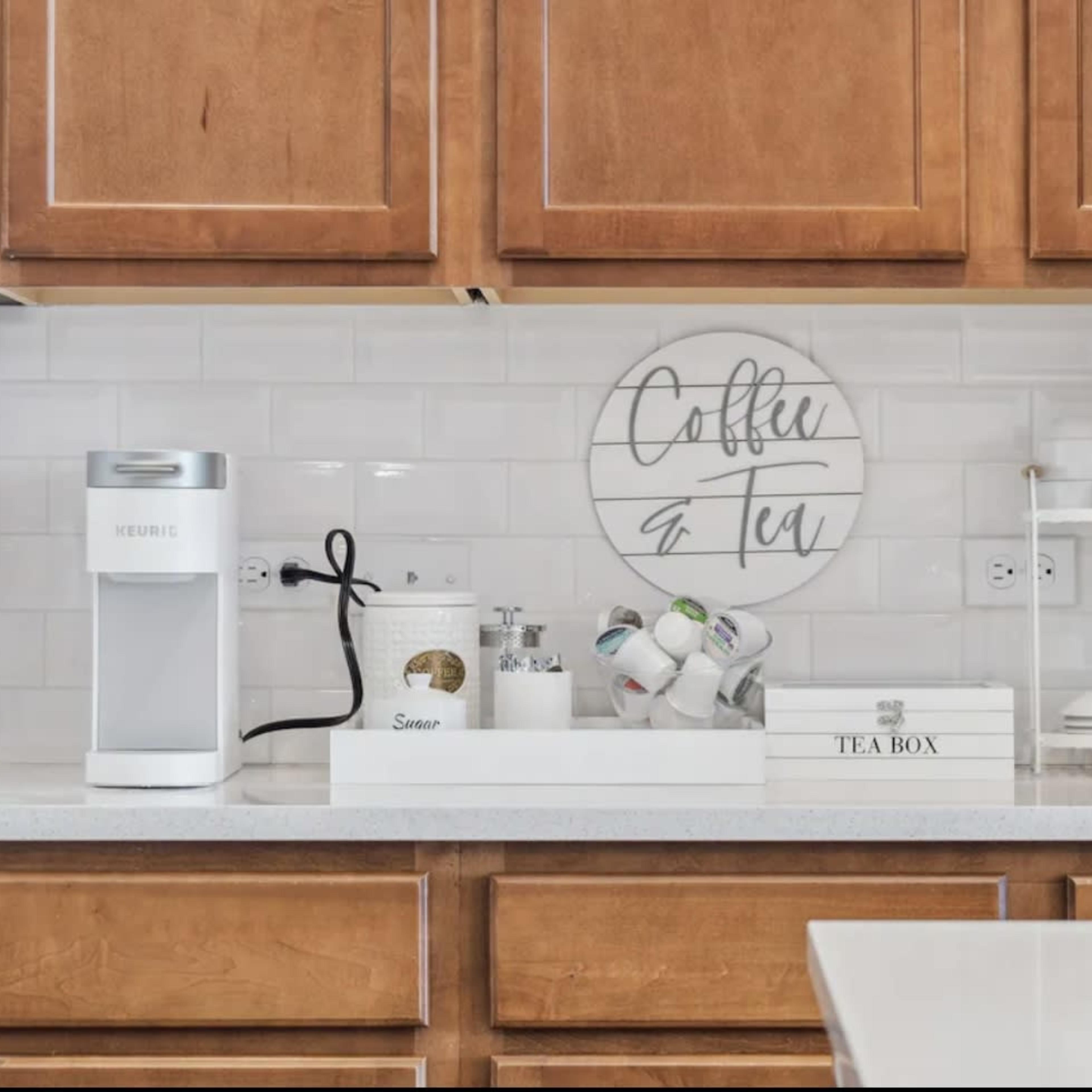 A kitchen counter with a coffee maker, tea kettle, decorative canisters, and a display of tea items against wooden cabinetry and a tiled backsplash.