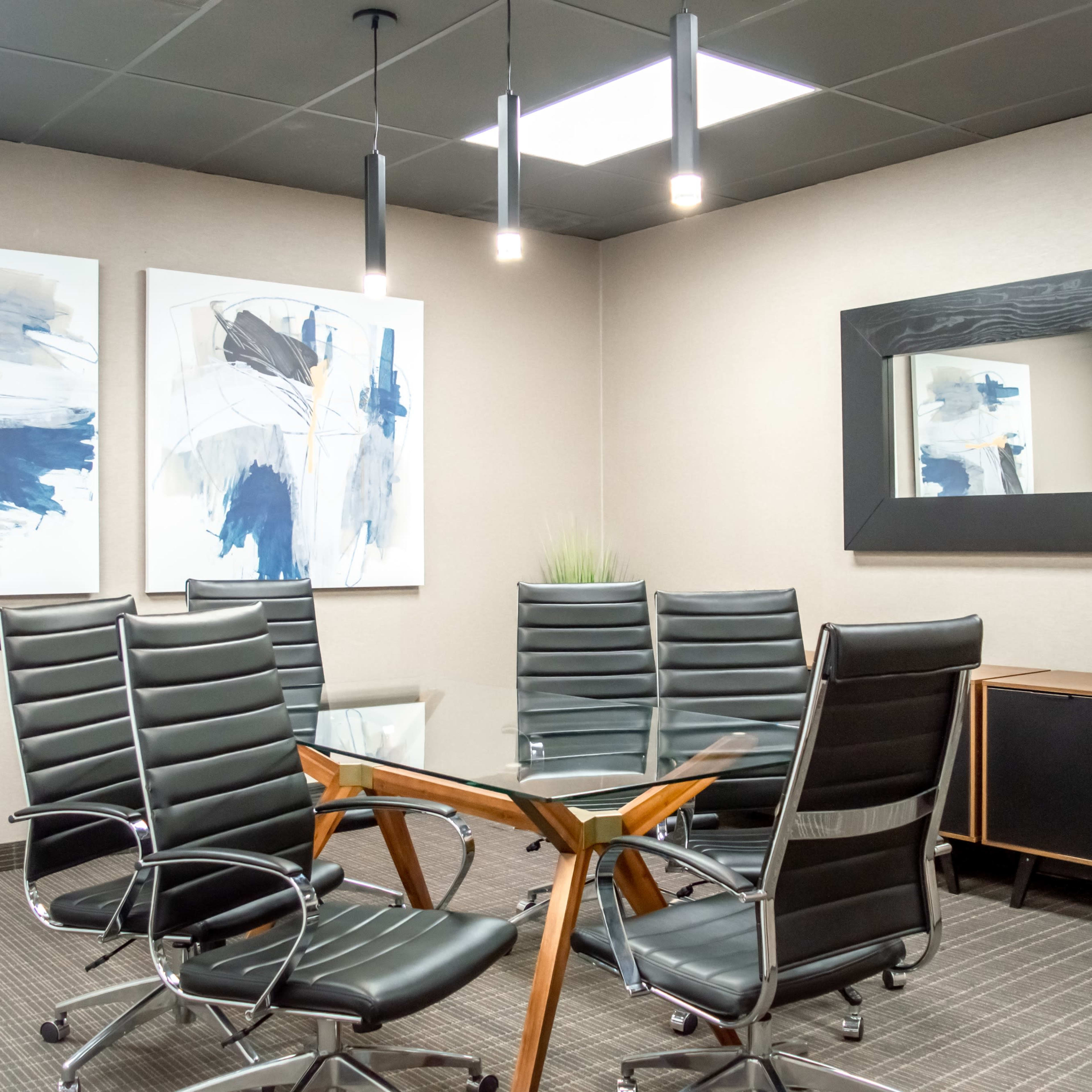 A modern conference room features a glass table surrounded by black leather chairs, with abstract artwork on the walls and a large mirror opposite the seating area.