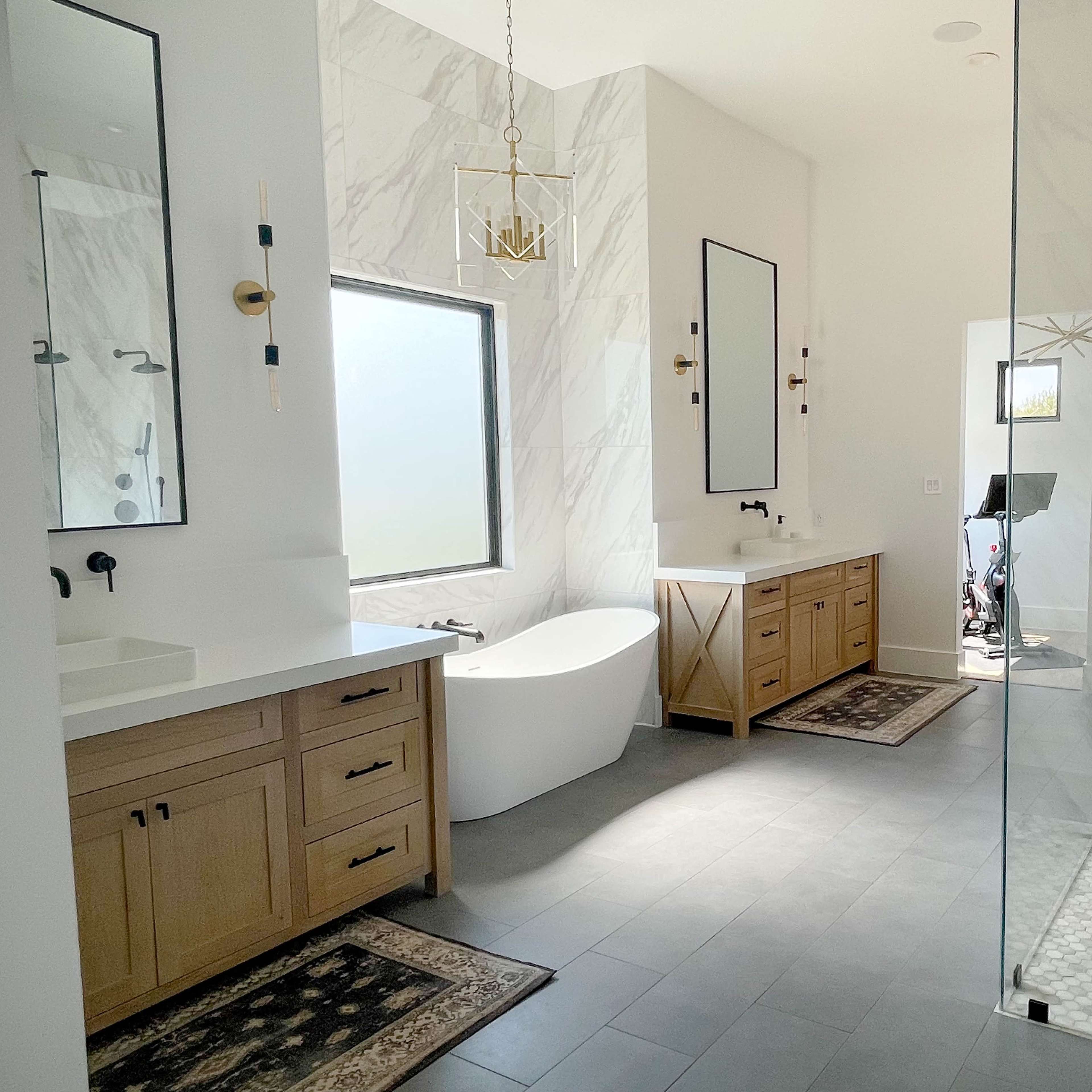 A modern bathroom featuring a freestanding bathtub, dual sinks with wooden vanities, a glass shower enclosure, and large mirrors against a marble backdrop.