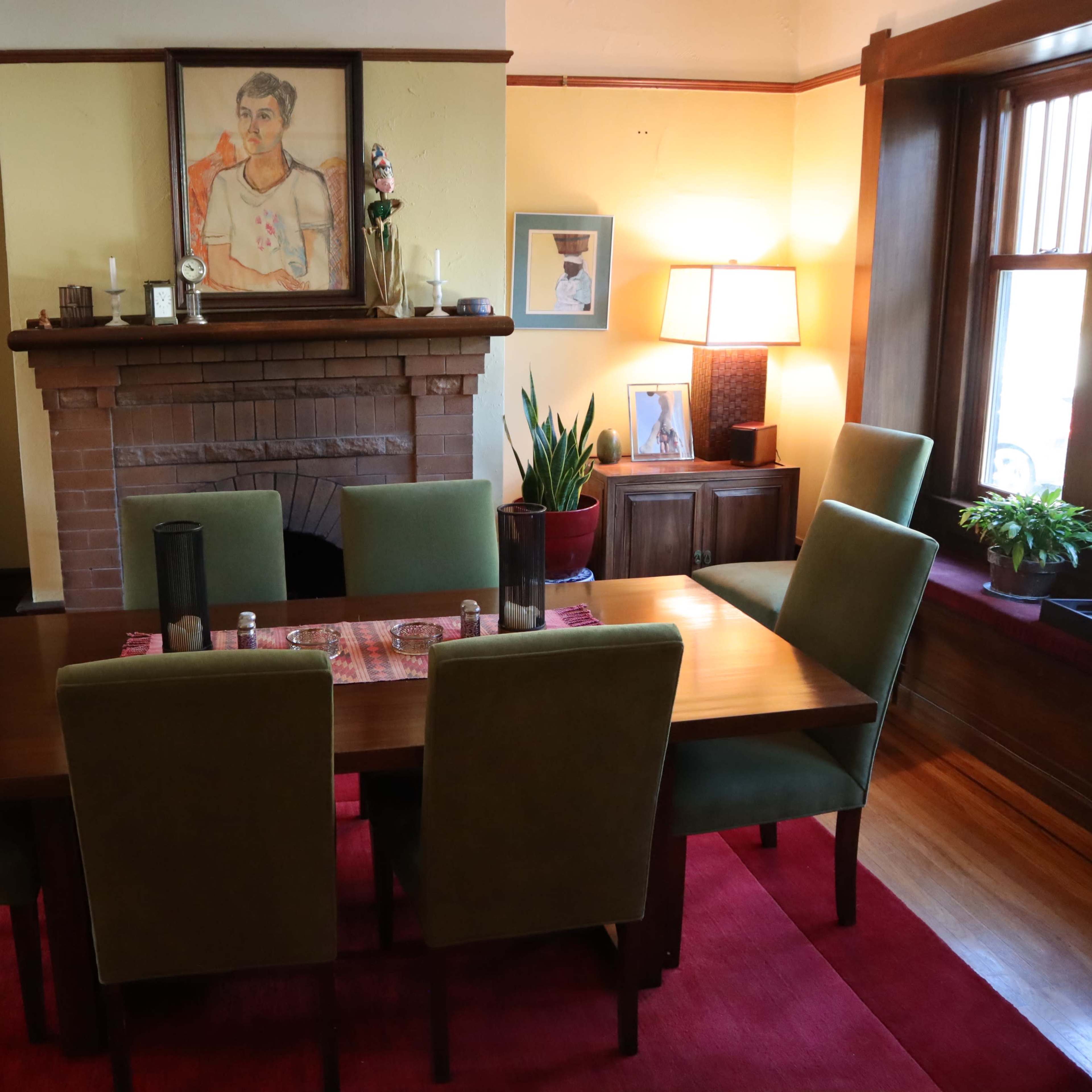 A dining room with a wooden table surrounded by green chairs, a fireplace with a painting above it, and windows allowing natural light.