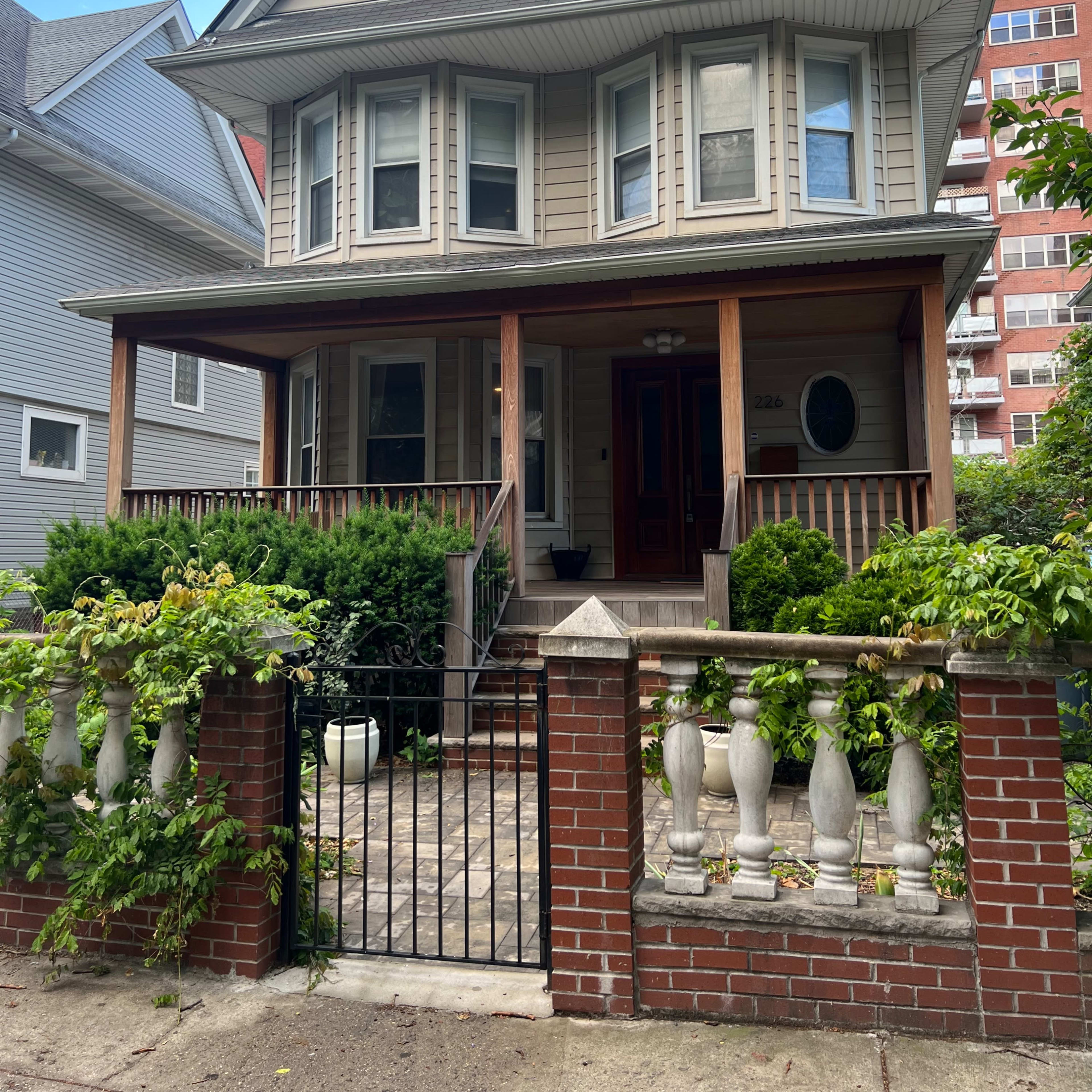 A three-story house with a front porch and decorative railing is situated behind a brick fence and surrounded by greenery.