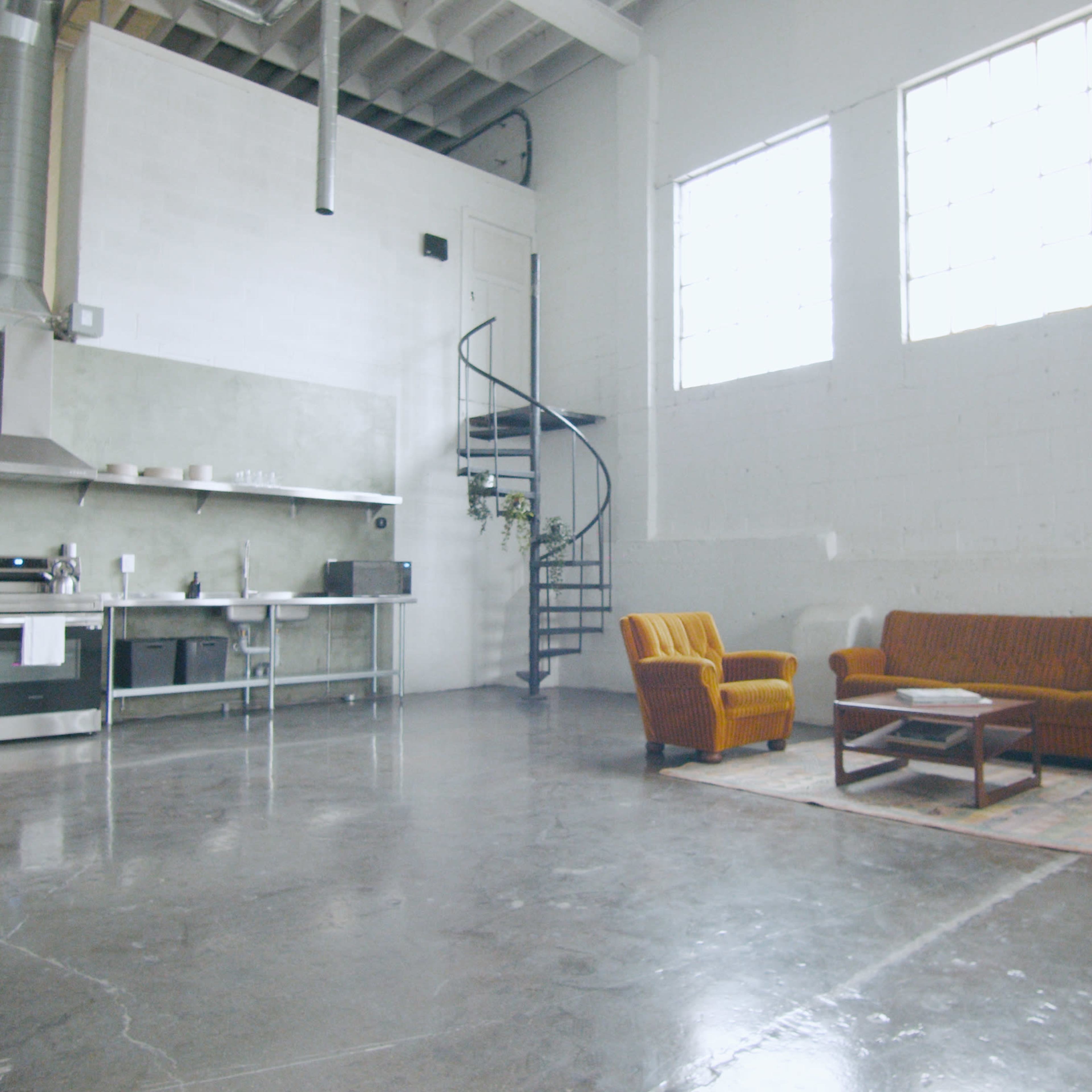 A spacious, industrial-style room featuring a kitchen area, a spiral staircase, and a seating area with a brown couch and armchair.