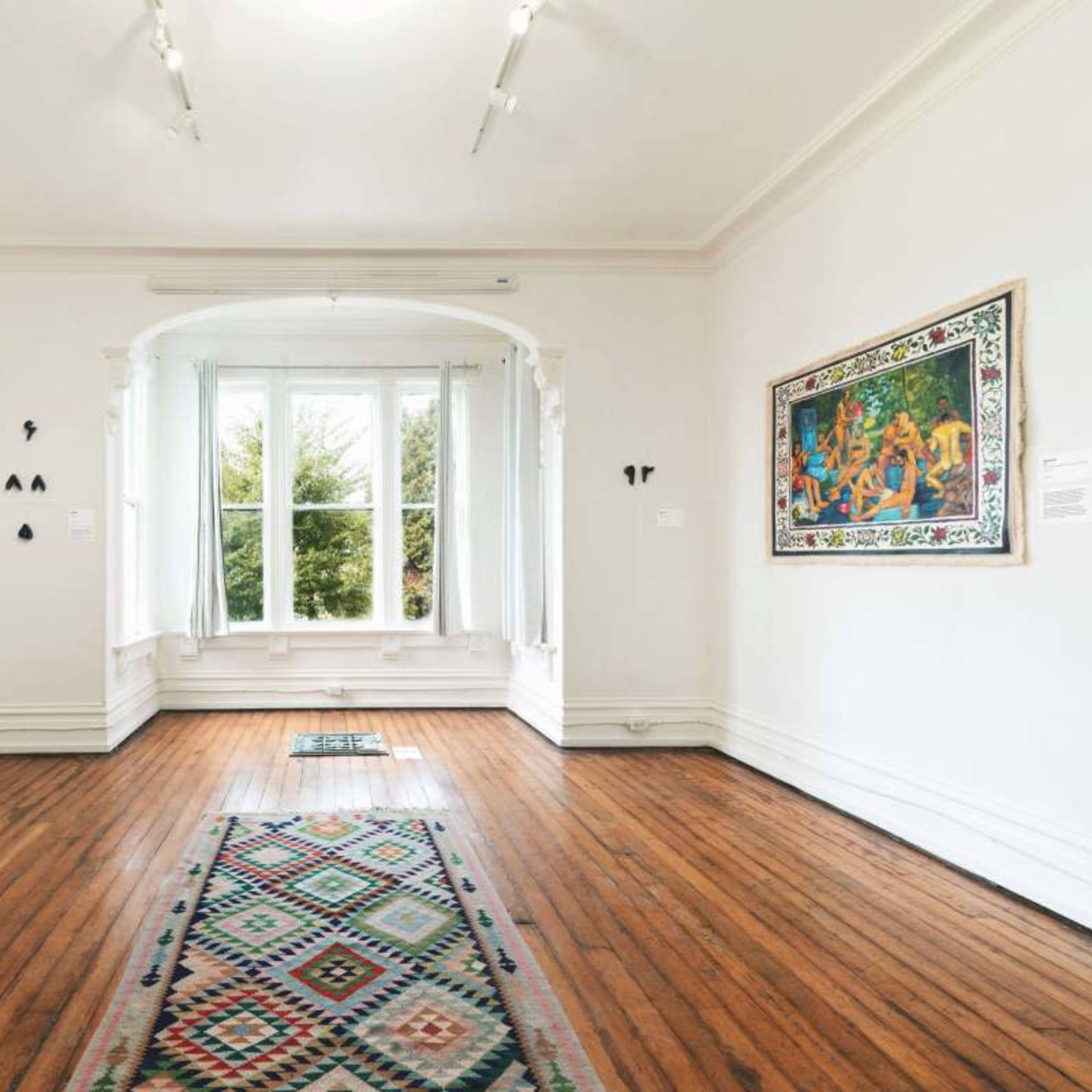 An art gallery with wooden floors, white walls, and several pieces of artwork displayed, along with a window that lets in natural light.