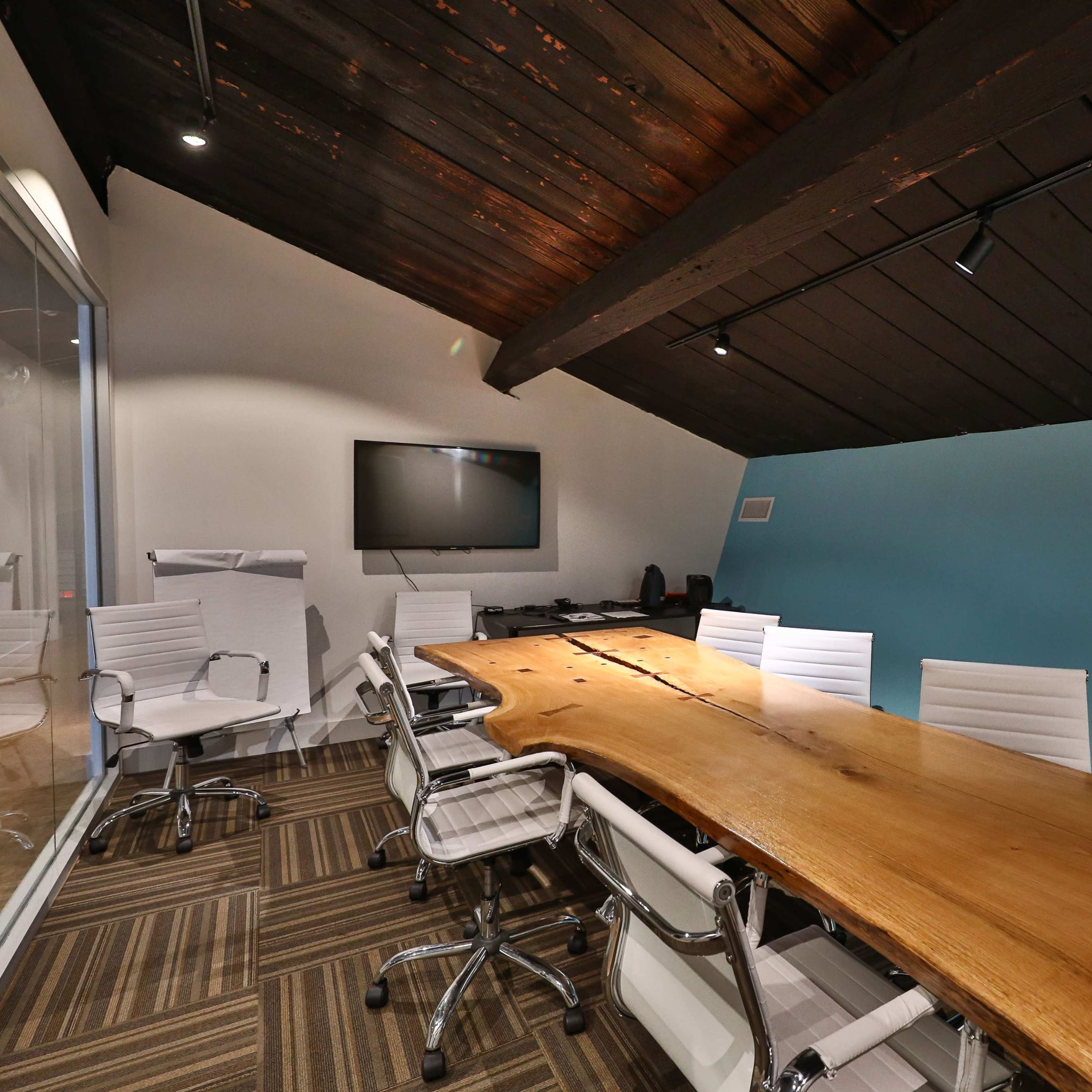 A modern conference room features a large wooden table surrounded by white chairs, with a blue accent wall and a television mounted on the wall.