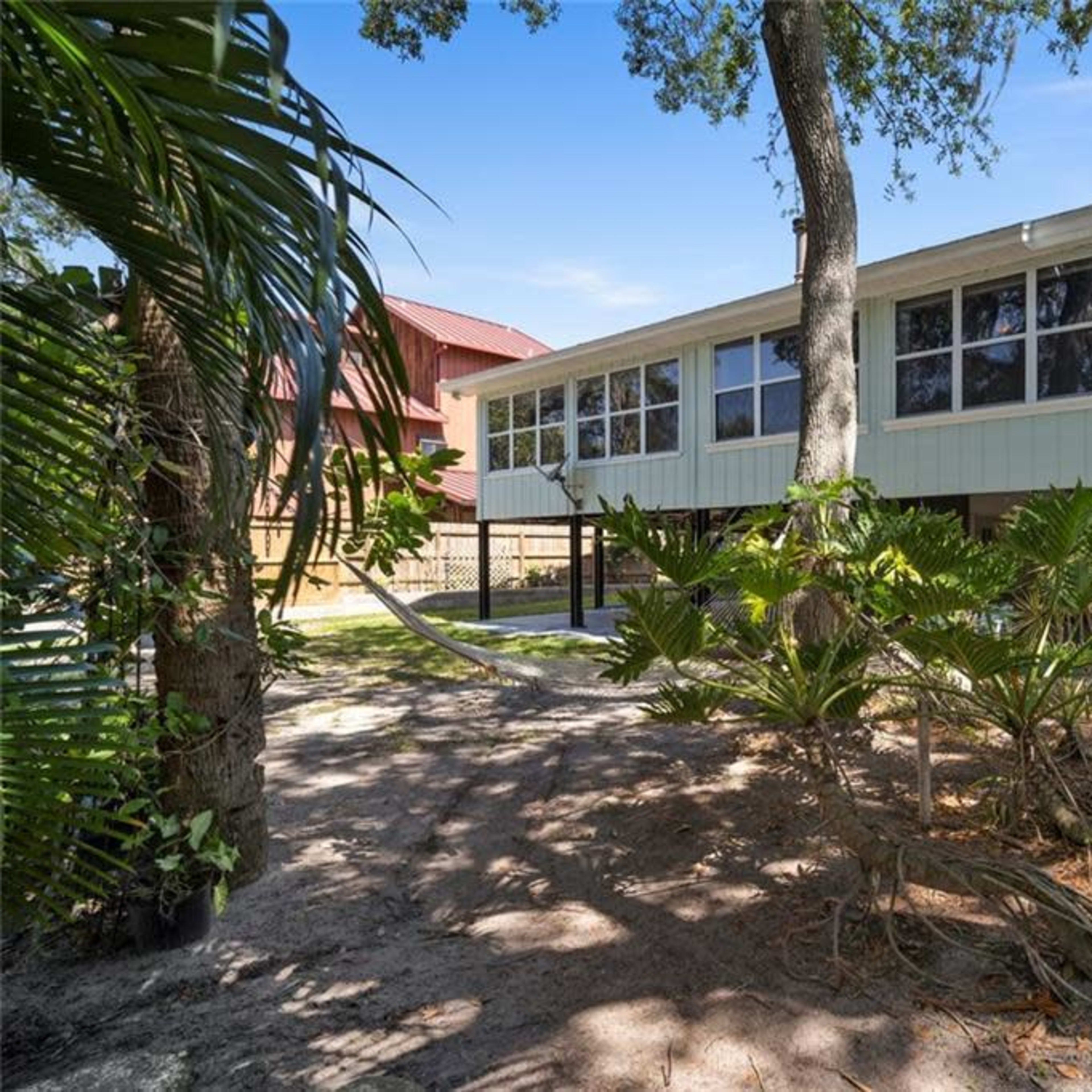 A house with a raised structure is surrounded by trees and tropical plants, with a sandy area beneath it.